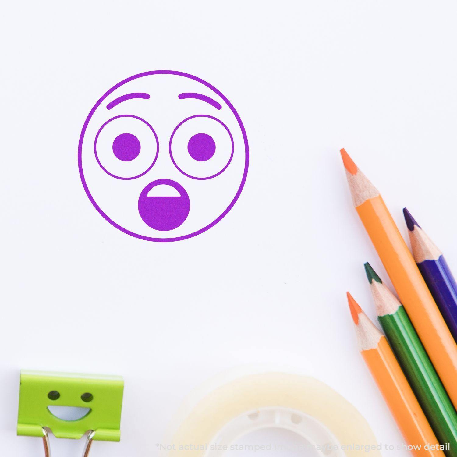 Round Surprised Smiley Rubber Stamp in purple ink on white paper, surrounded by colored pencils, a green clip, and tape.