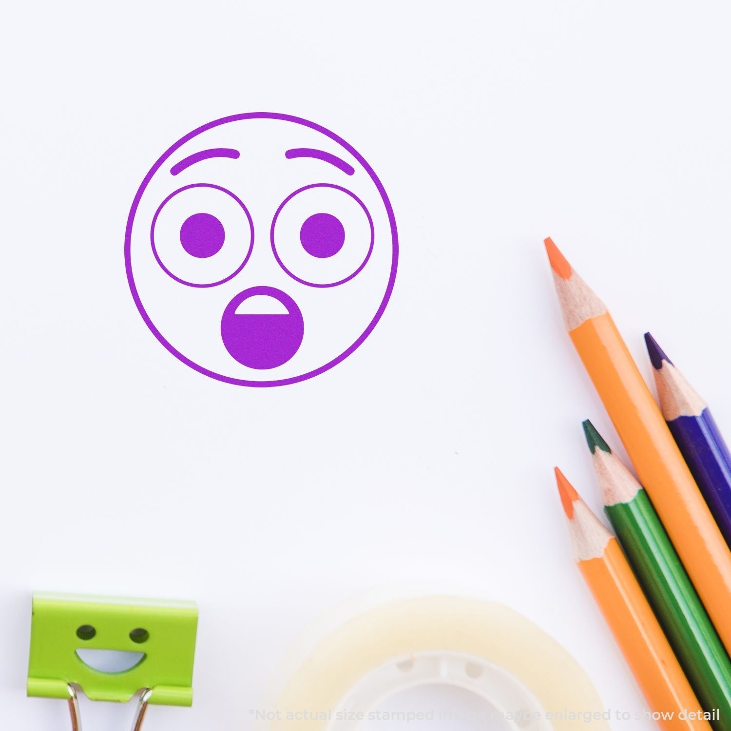 Self Inking Round Surprised Smiley Stamp in purple ink on white paper, surrounded by colored pencils, a green clip, and tape.