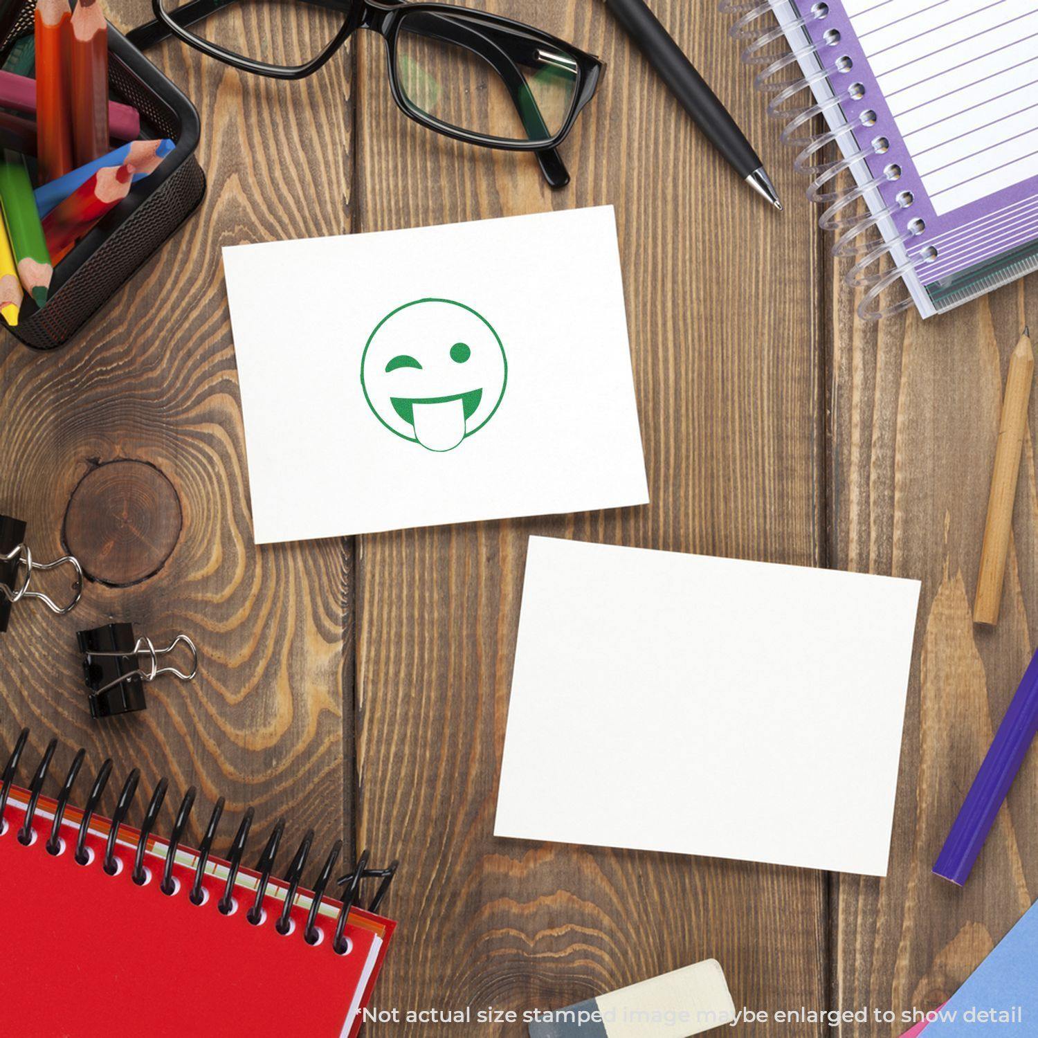 Round Tongue Out Smiley Rubber Stamp on a white card, surrounded by office supplies on a wooden desk.