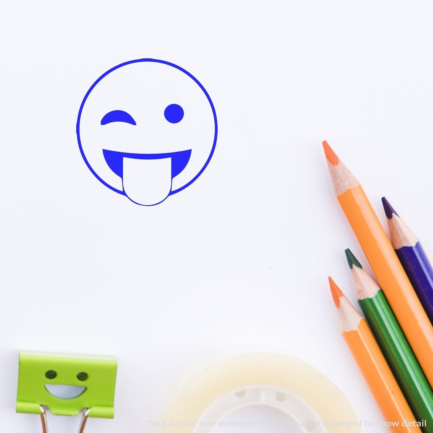 Self Inking Round Tongue out smiley Stamp imprint on paper, surrounded by colorful pencils, a green clip, and tape.