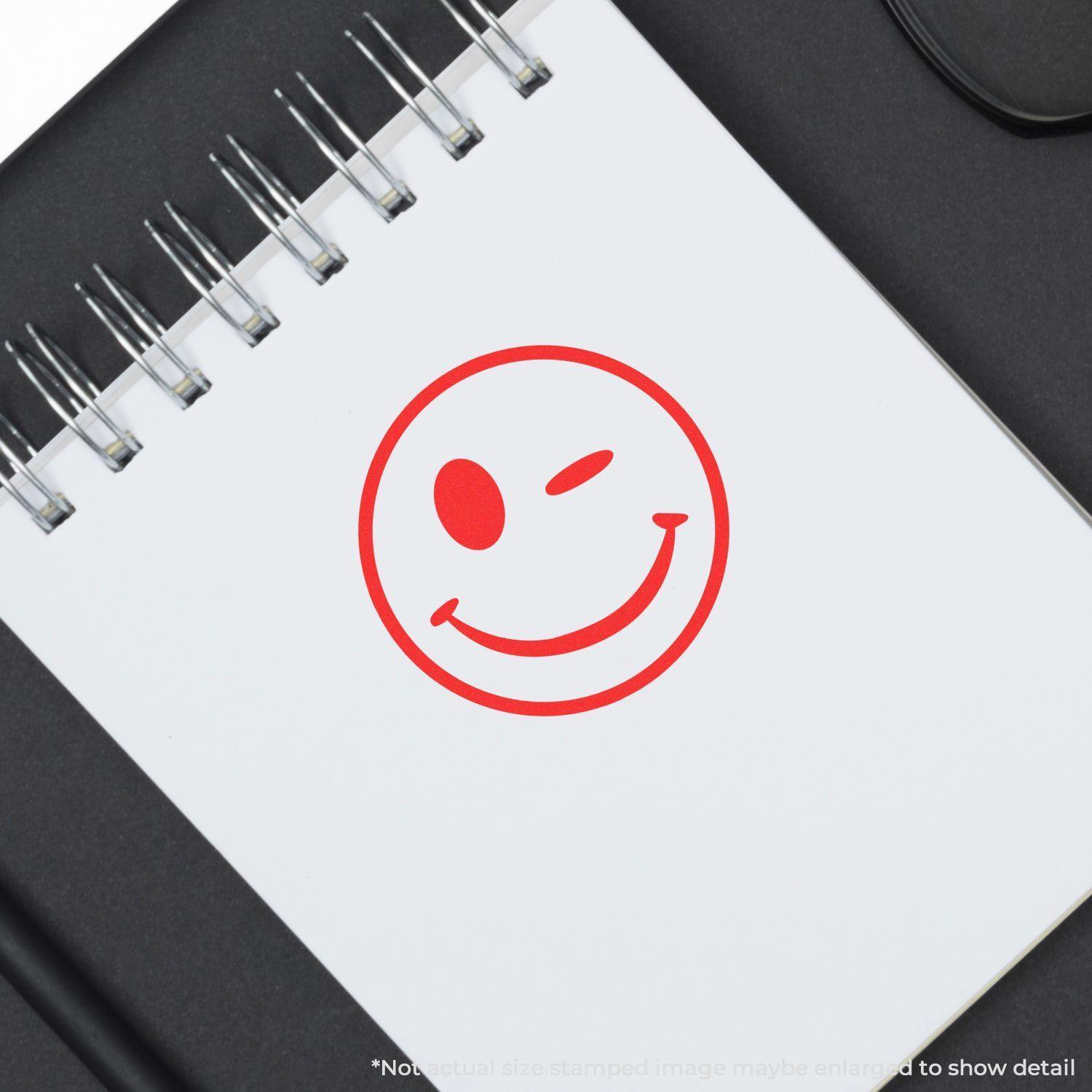 Round Winking Smiley Rubber Stamp in red ink on a white notepad with a spiral binding, placed on a black surface.