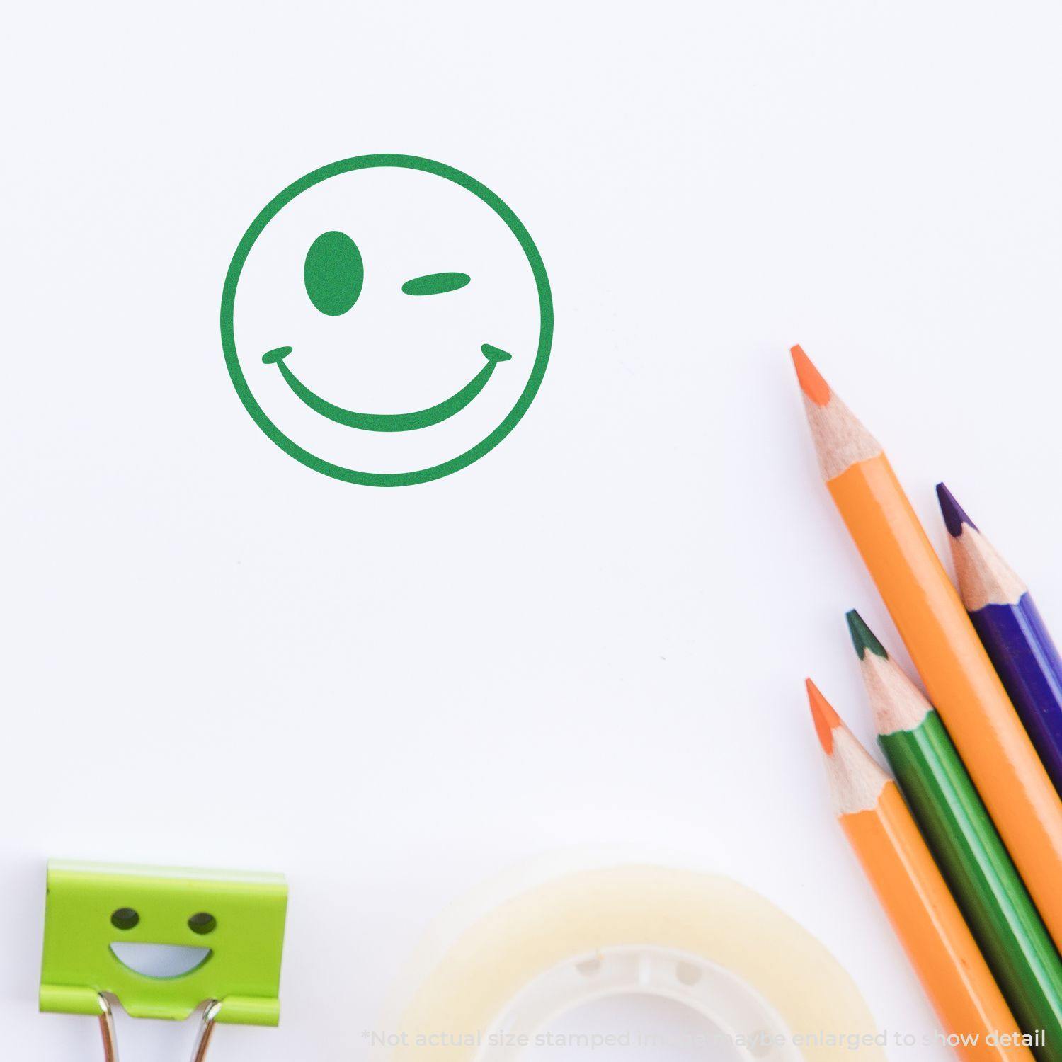 Round Winking Smiley Rubber Stamp in green ink on white paper, surrounded by colored pencils, a smiley clip, and tape.