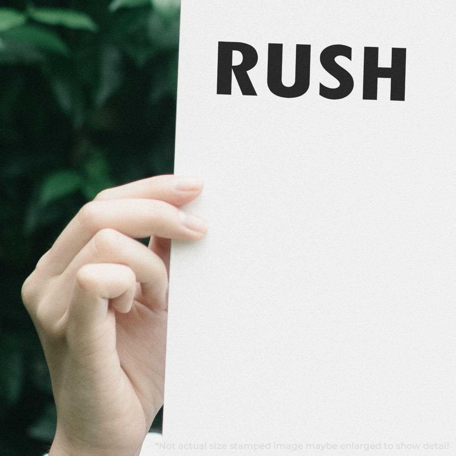 Hand holding a paper stamped with RUSH using the Large Pre-Inked Rush Stamp, with a blurred green background.