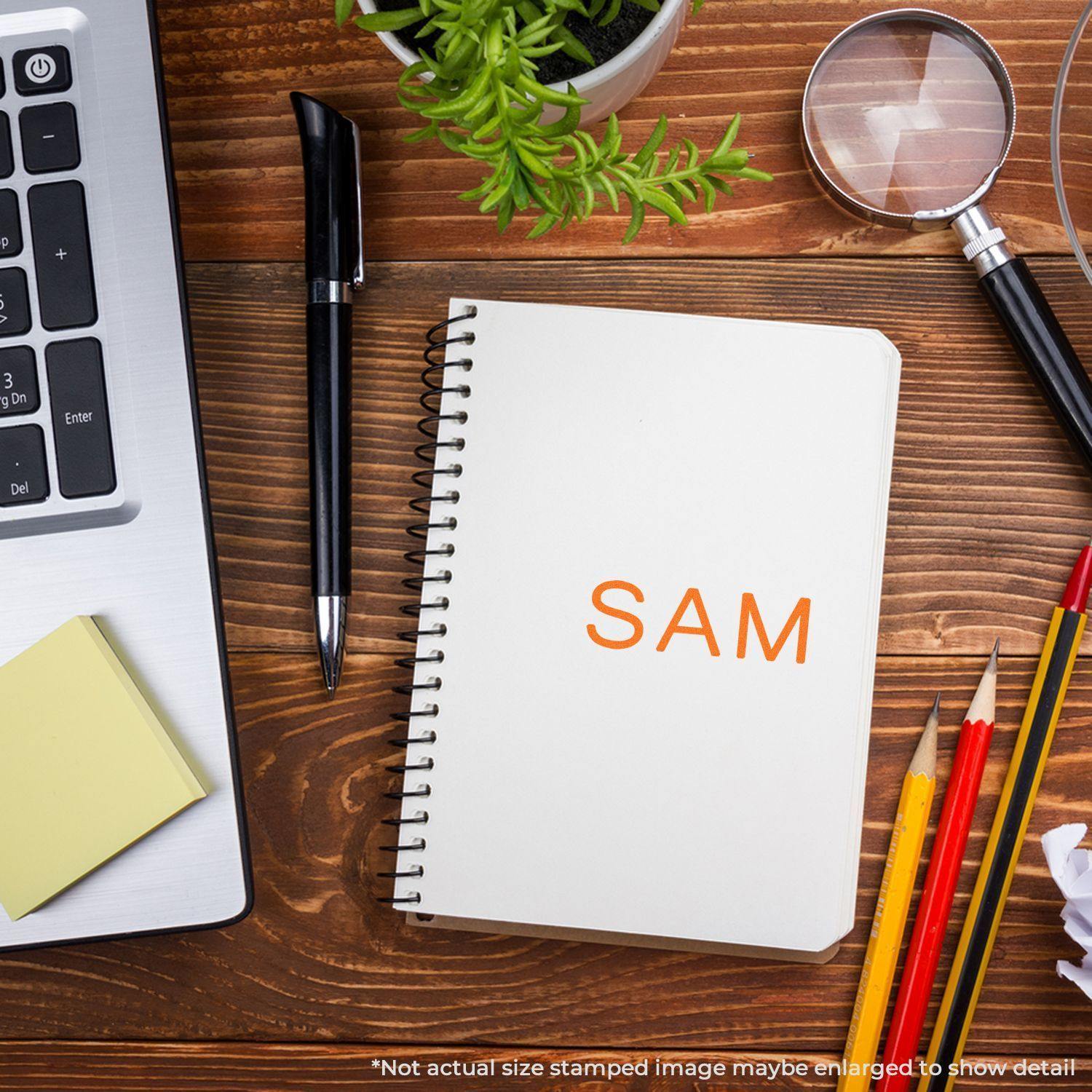 Large Pre-Inked SAM Stamp used on a notebook, surrounded by office supplies including a laptop, pen, magnifying glass, and plant.