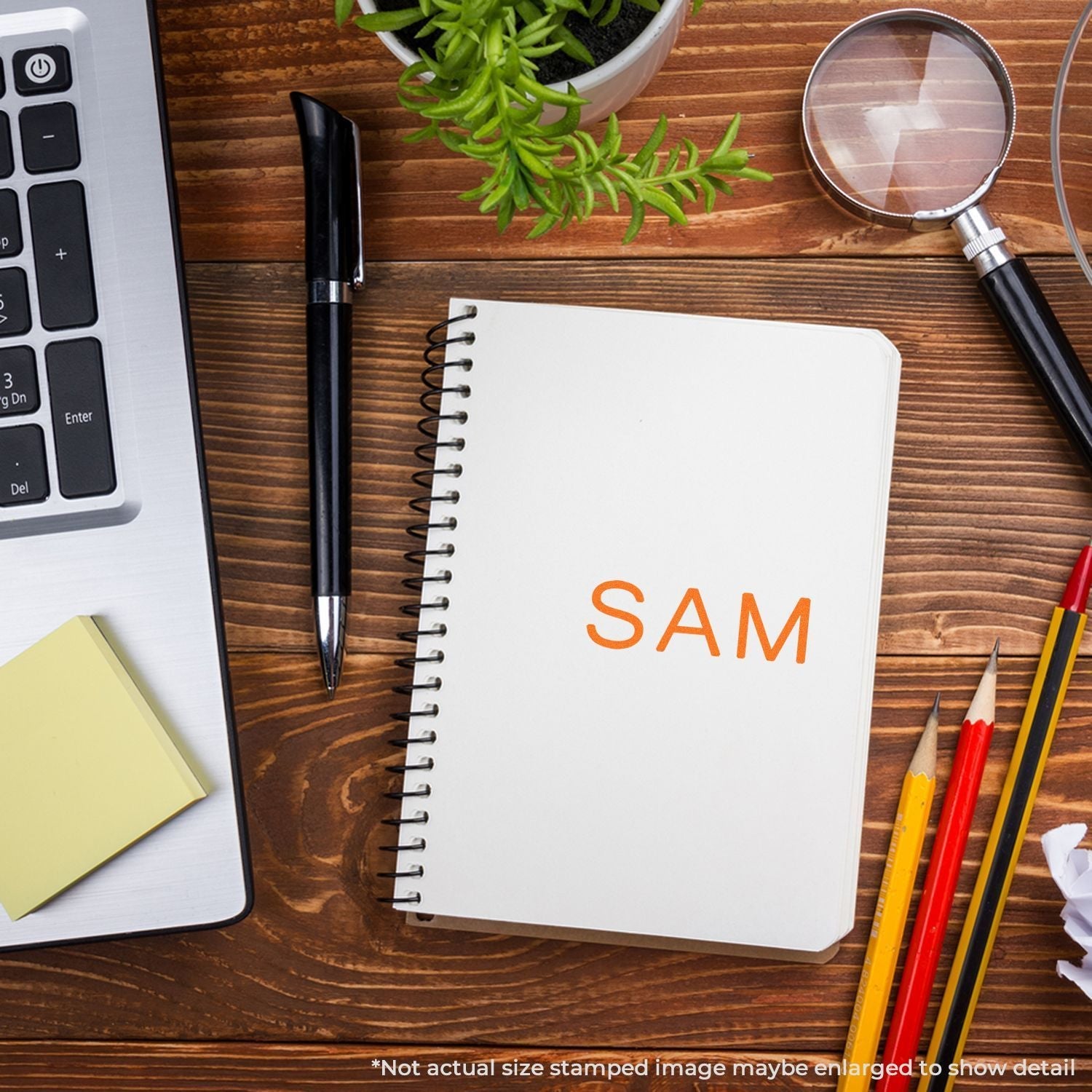 Self Inking SAM Stamp used on a notebook, placed on a wooden desk with a laptop, plant, magnifying glass, pen, sticky notes, and pencils.