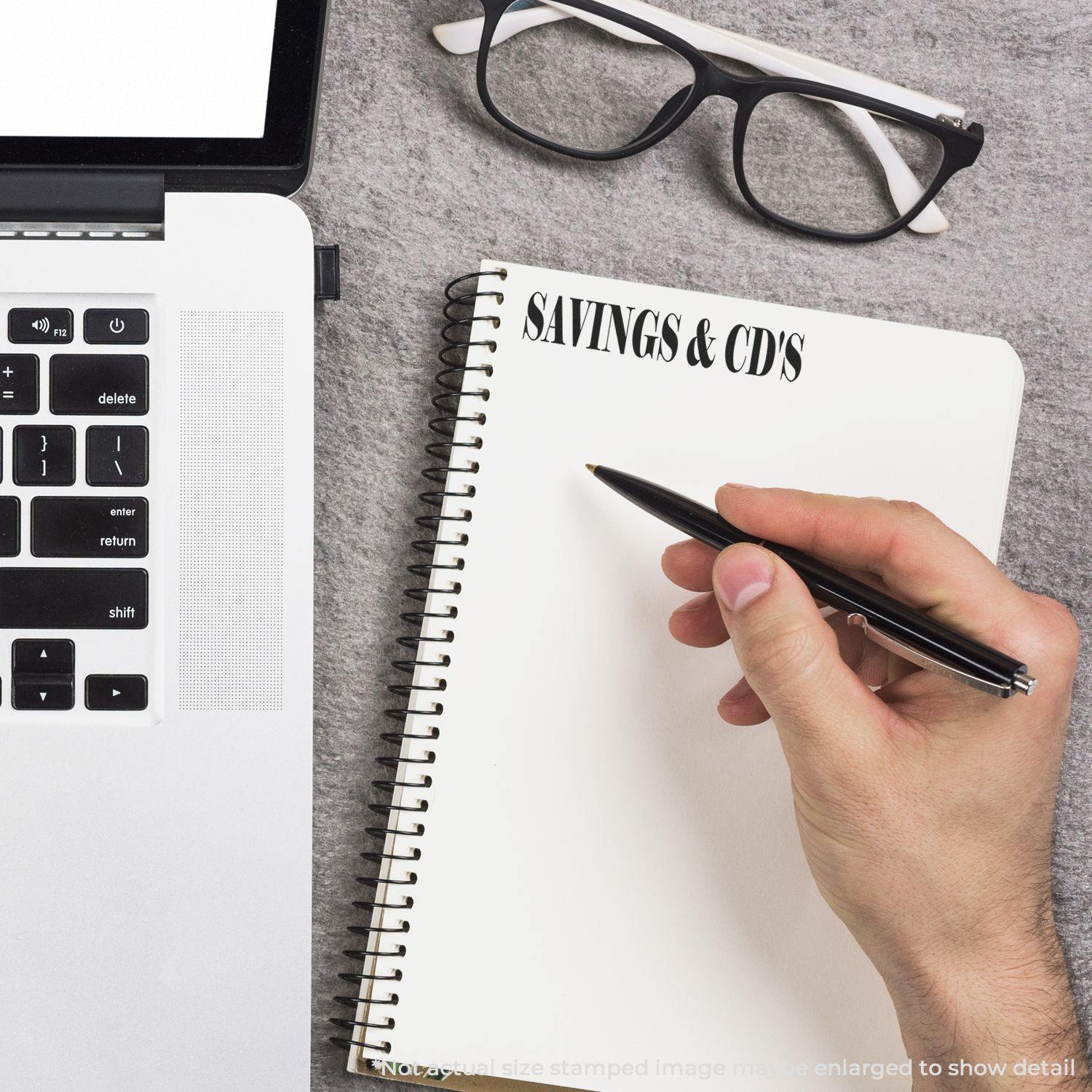 Hand writing in a notebook labeled SAVINGS & CDS next to a laptop and glasses on a gray surface.