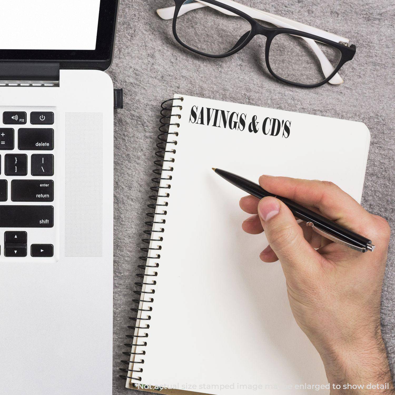 Person writing in a notebook stamped with SAVINGS & CD'S using the Large Savings & CD's Rubber Stamp, next to a laptop and glasses.