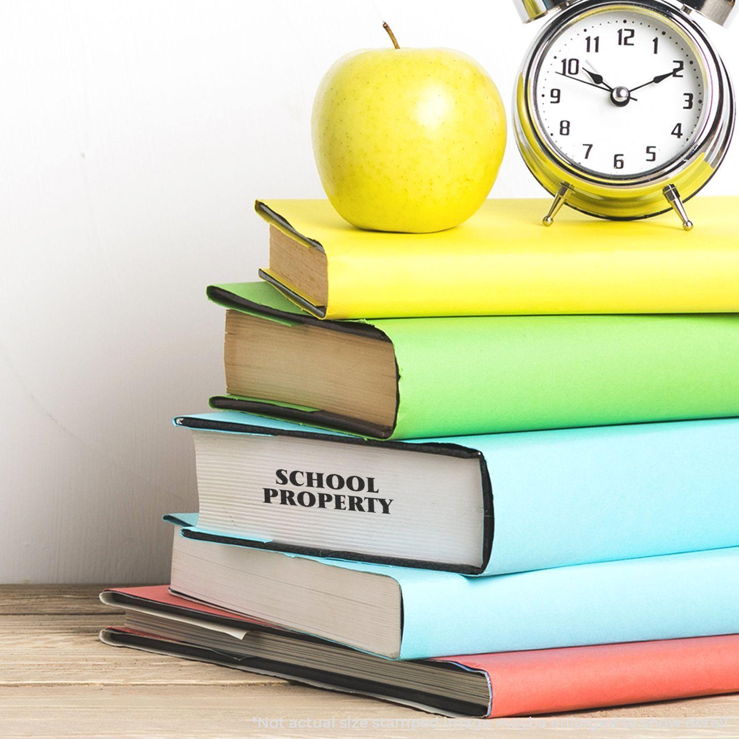 Stack of colorful books with a Self Inking School Property Stamp, a green apple, and a clock on a wooden surface.