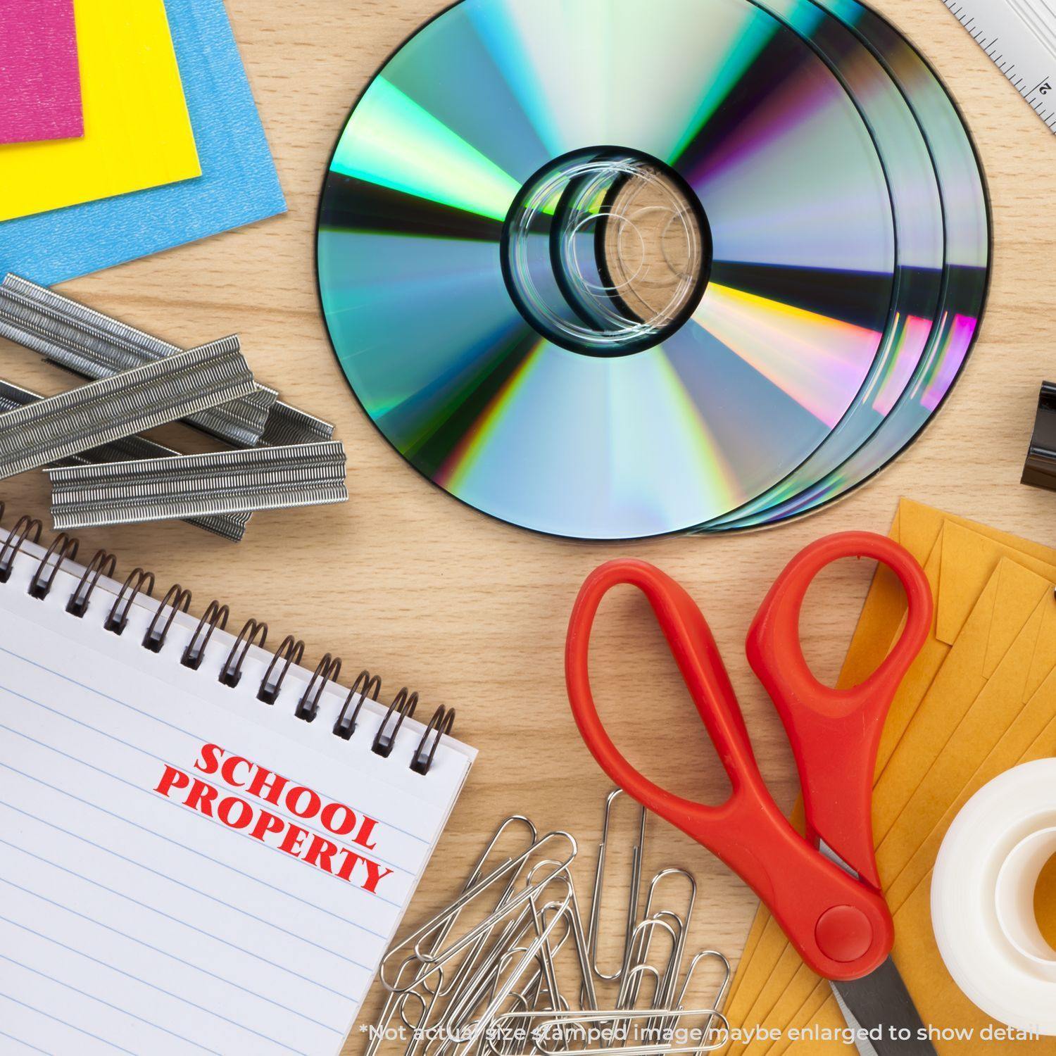 Slim Pre-Inked School Property Stamp on a notepad surrounded by CDs, red scissors, paper clips, and colorful paper on a desk.