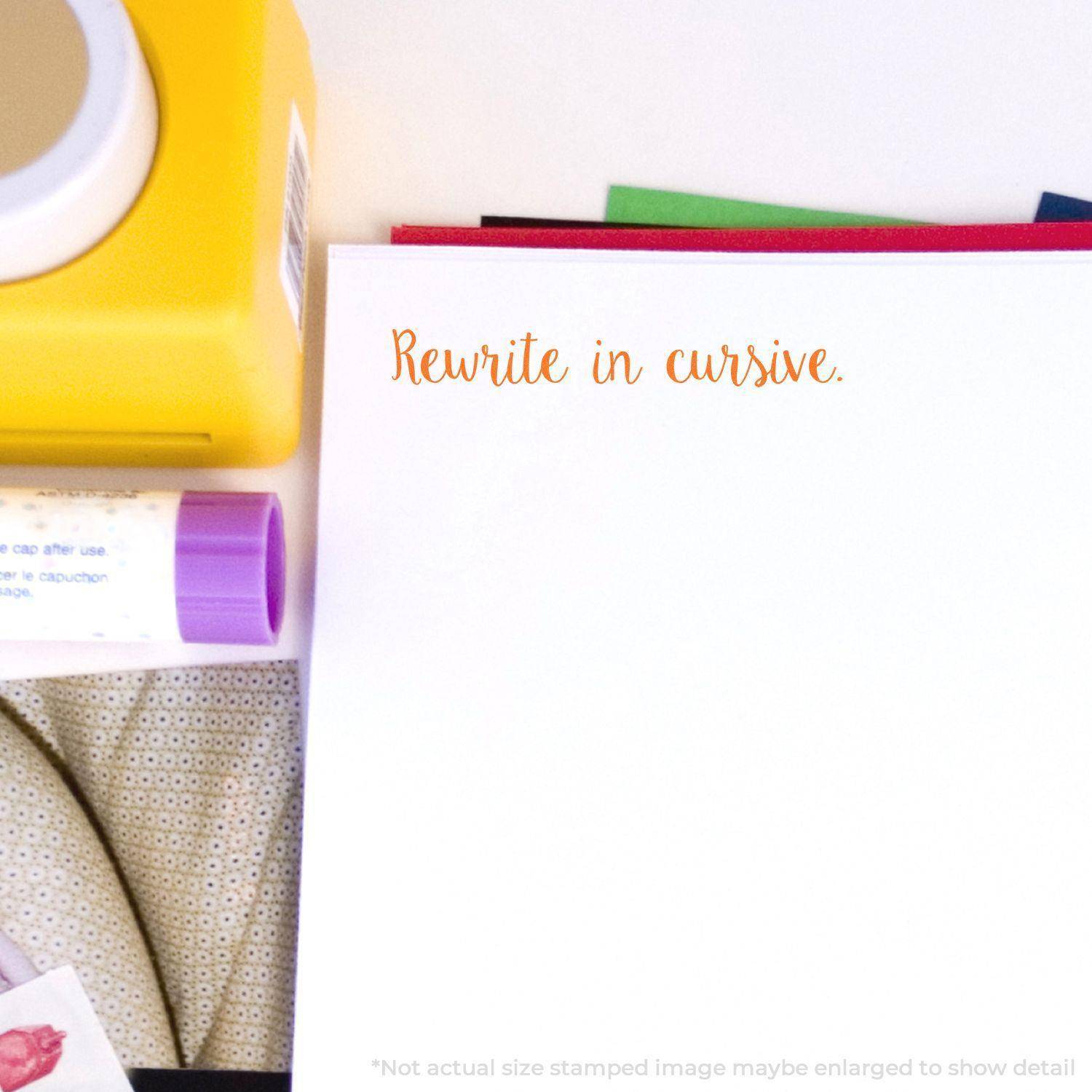 A Large Script Rewrite in Cursive Rubber Stamp is used on white paper, showing the text Rewrite in cursive in orange ink. Office supplies are nearby.