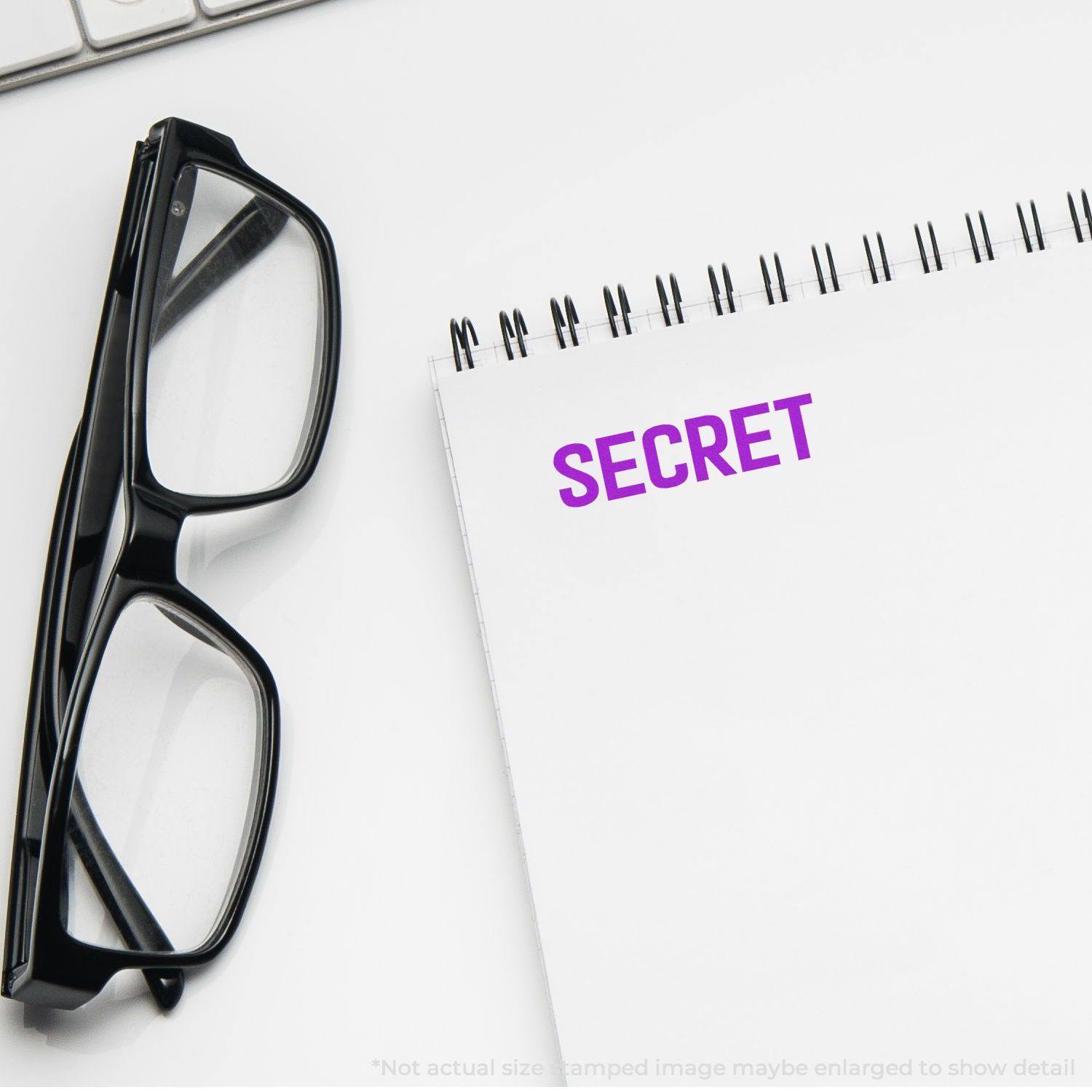 Self Inking Secret Stamp imprinting SECRET in purple on a notepad, with black glasses and a keyboard nearby.