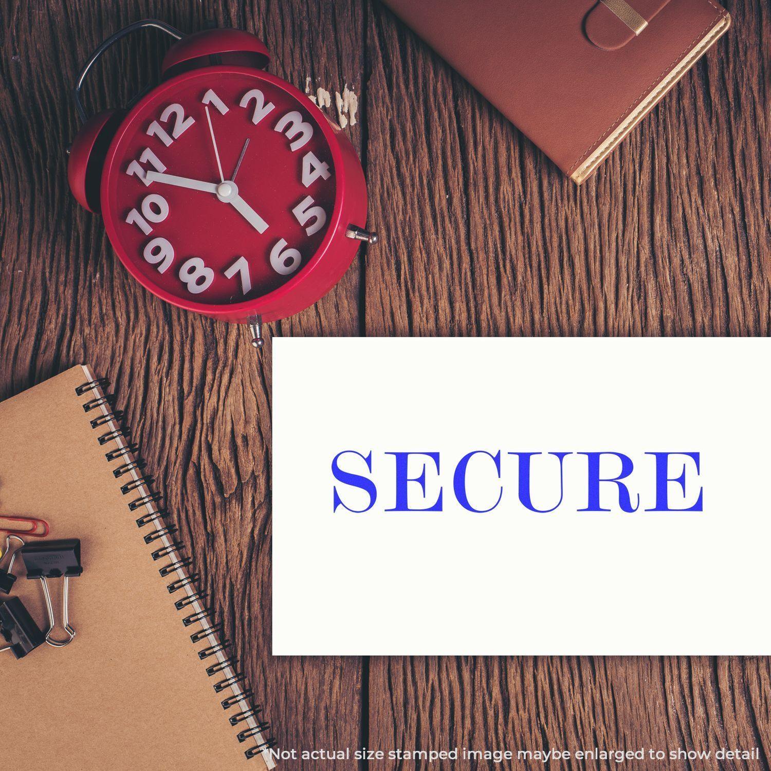 Large Pre-Inked Secure Stamp used on a white paper, placed on a wooden desk with a red clock, notebook, and office supplies.