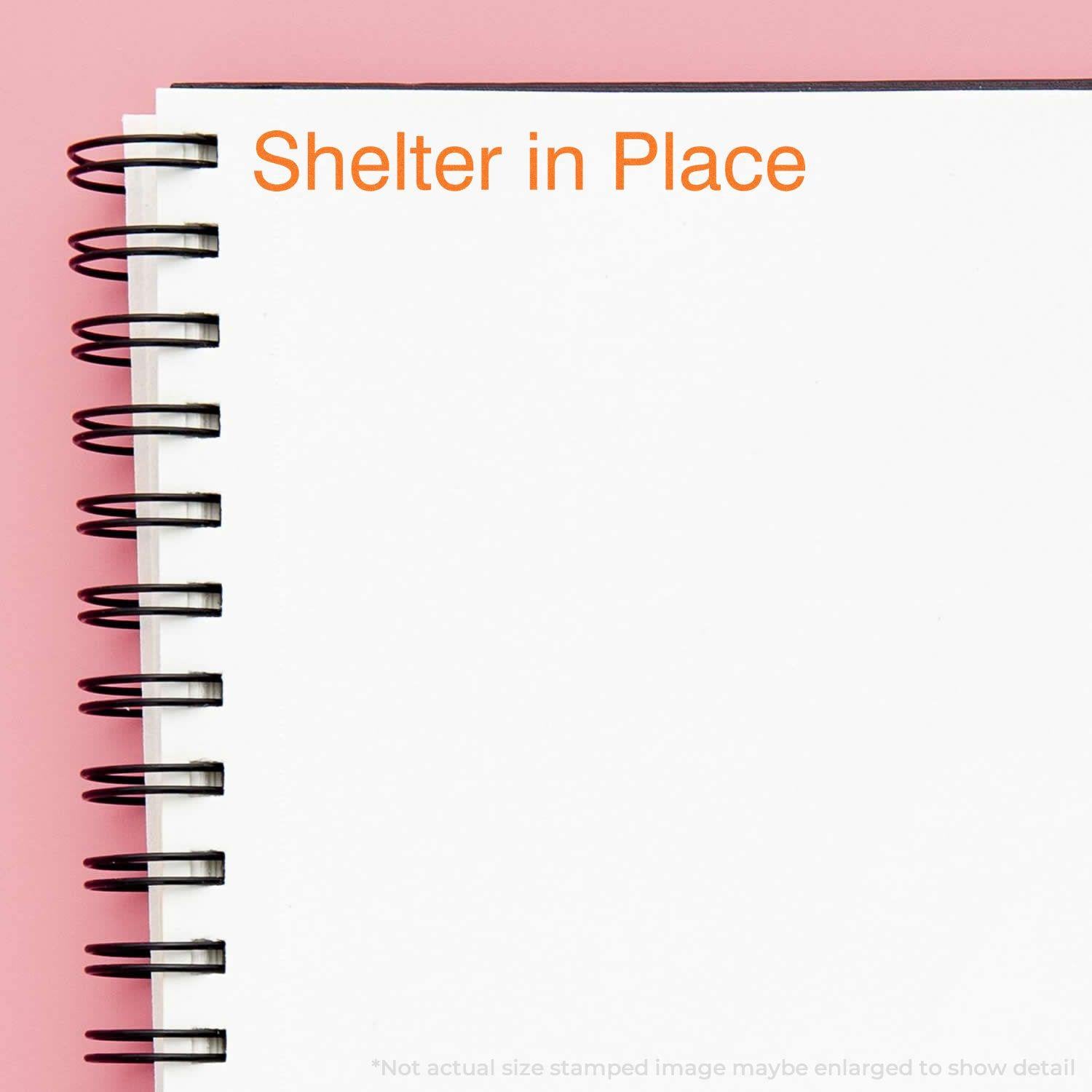 Shelter in Place rubber stamp impression in orange ink on a white spiral notebook against a pink background.