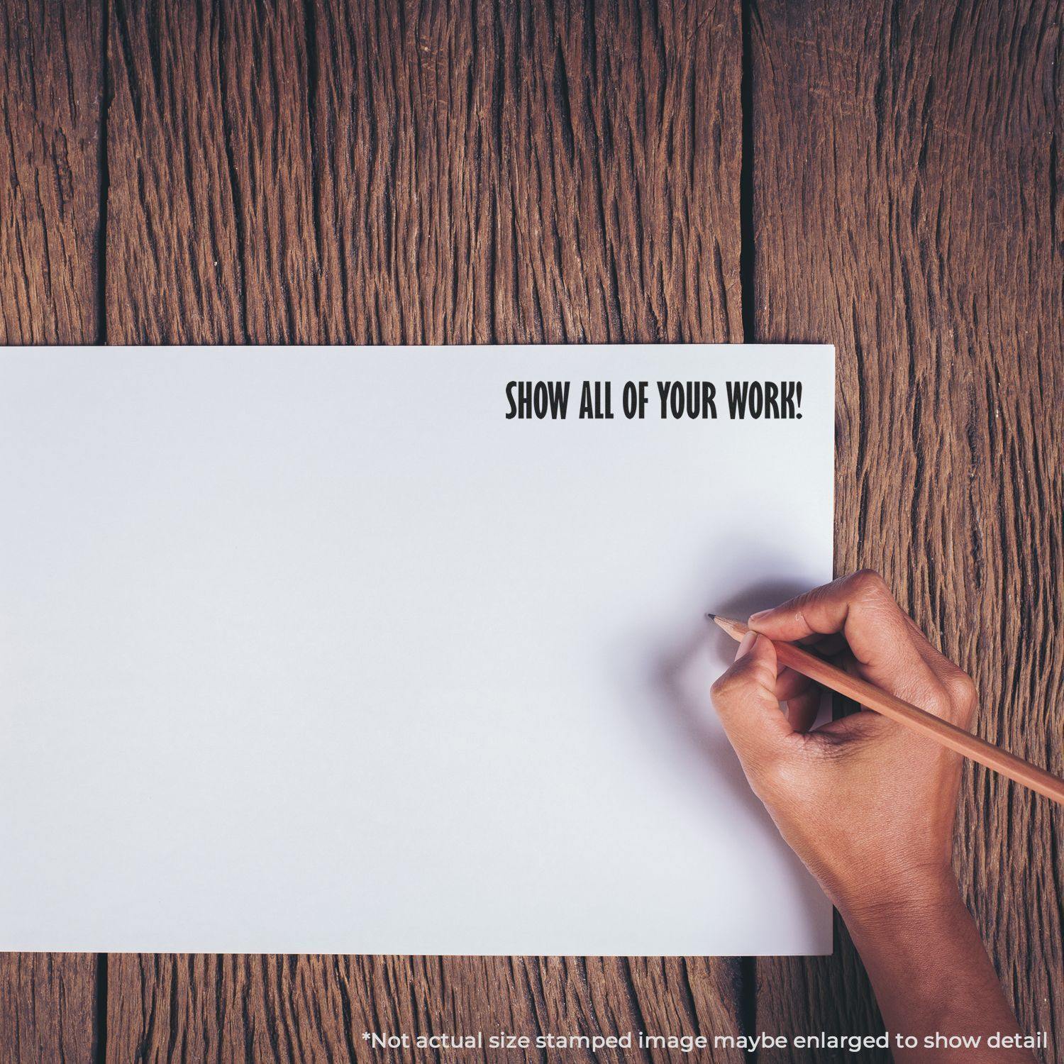 Hand holding pencil near paper with Show All Of Your Work rubber stamp text on wooden surface.