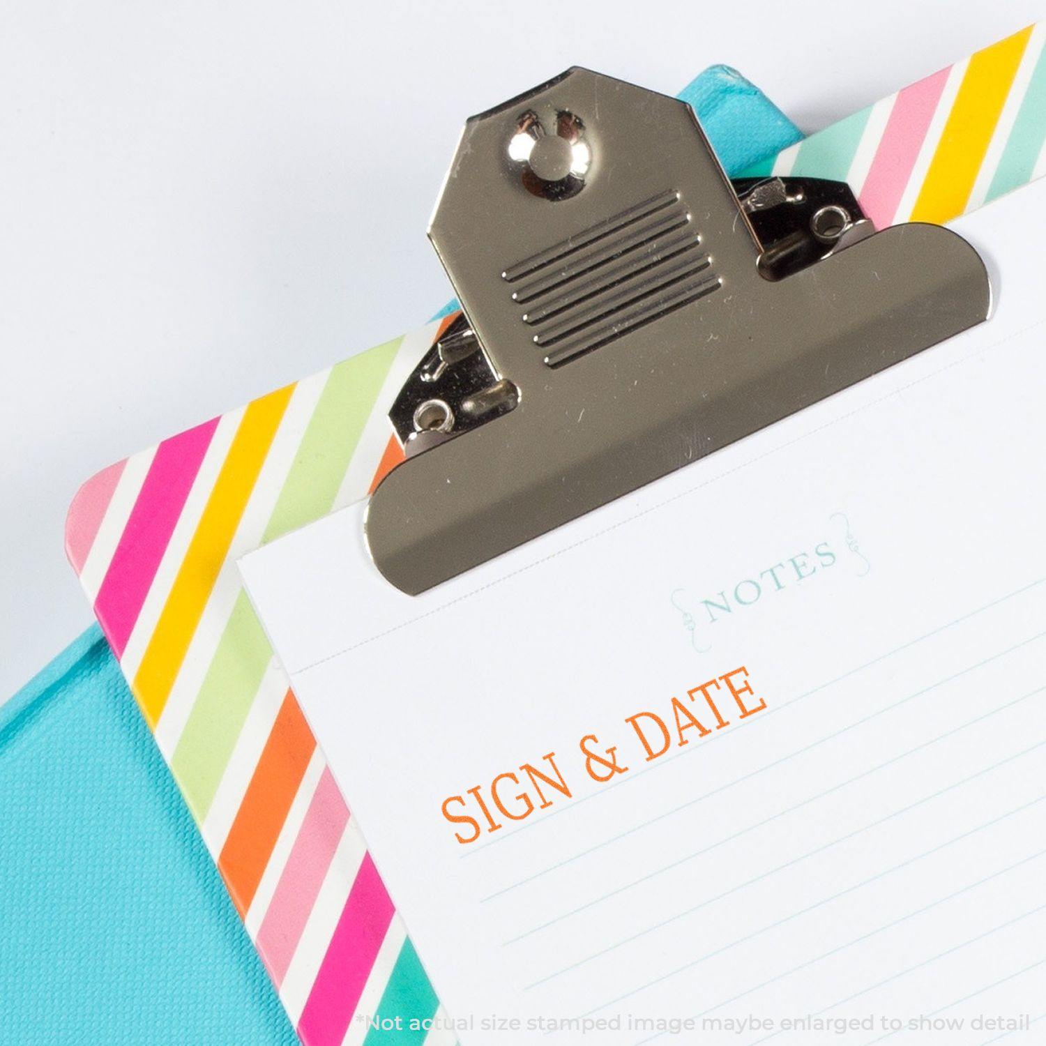 Self Inking Sign & Date Stamp in use on a colorful clipboard with a Notes page, showing the stamped text SIGN & DATE in orange.