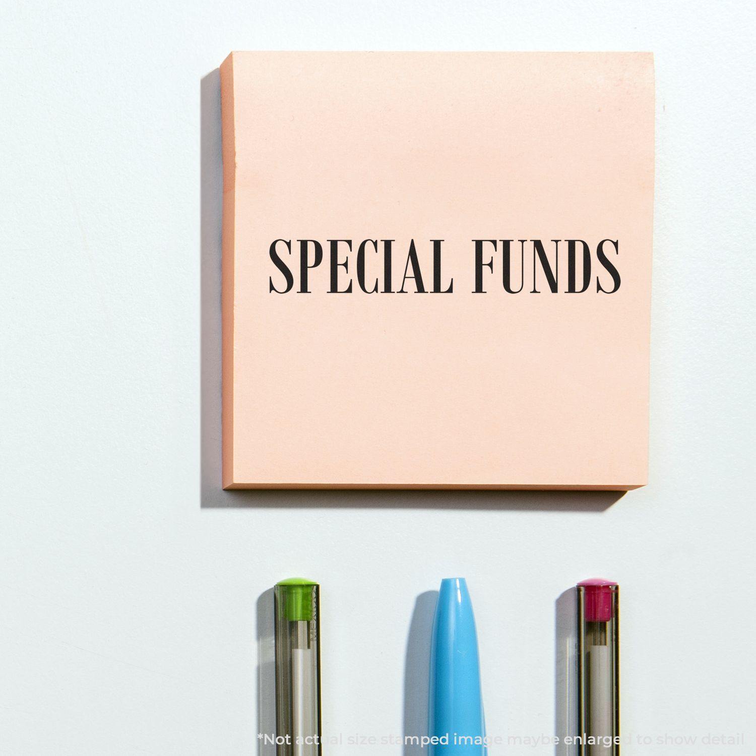 Large Pre-Inked Special Funds Stamp used on a pink sticky note, with three pens below it on a white surface.