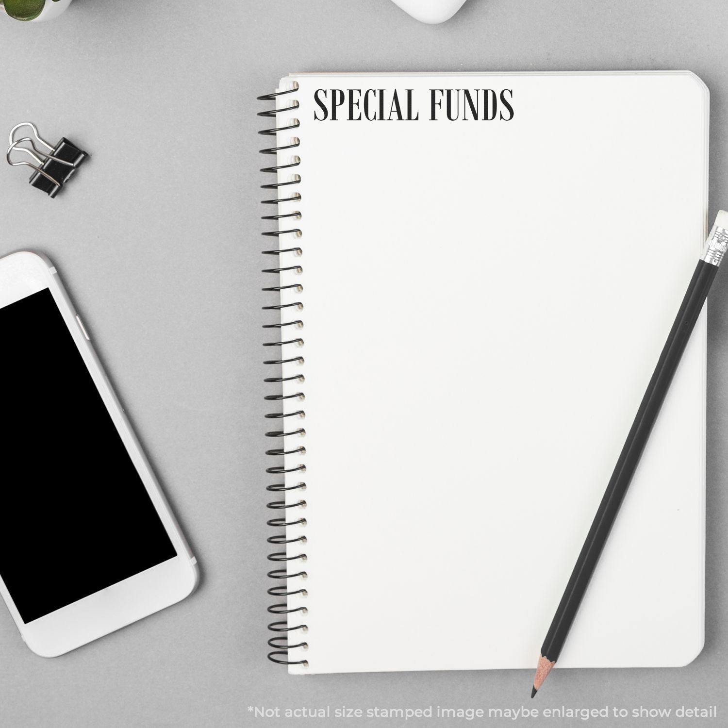 Large Pre-Inked Special Funds Stamp used on a spiral notebook, with a pencil, smartphone, and paper clip nearby on a gray surface.