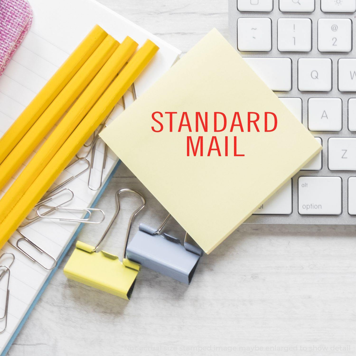 Large Self Inking Standard Mail Stacked Stamp on a yellow sticky note near a keyboard, pencils, and paper clips.