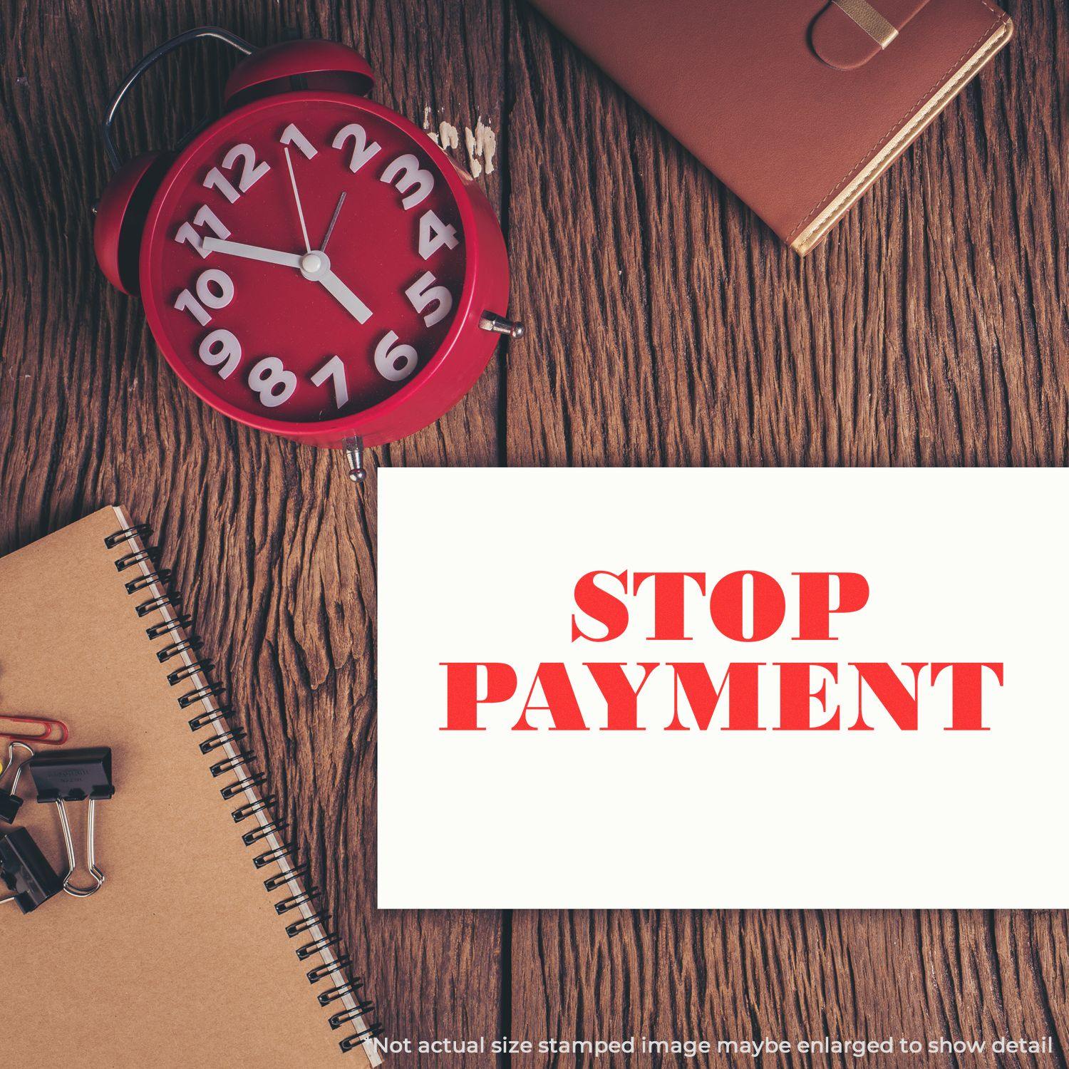 Large Pre-Inked Stop Payment Stamp used on a white paper, placed on a wooden desk with a red clock, notebook, and office supplies.