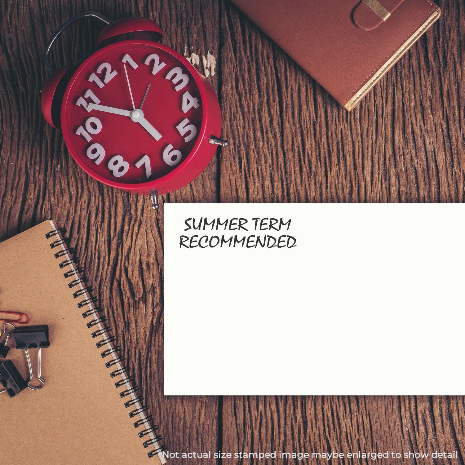 Self Inking Summer Term Recommended Stamp on a white paper, surrounded by a red clock, brown notebook, and office supplies on a wooden desk.