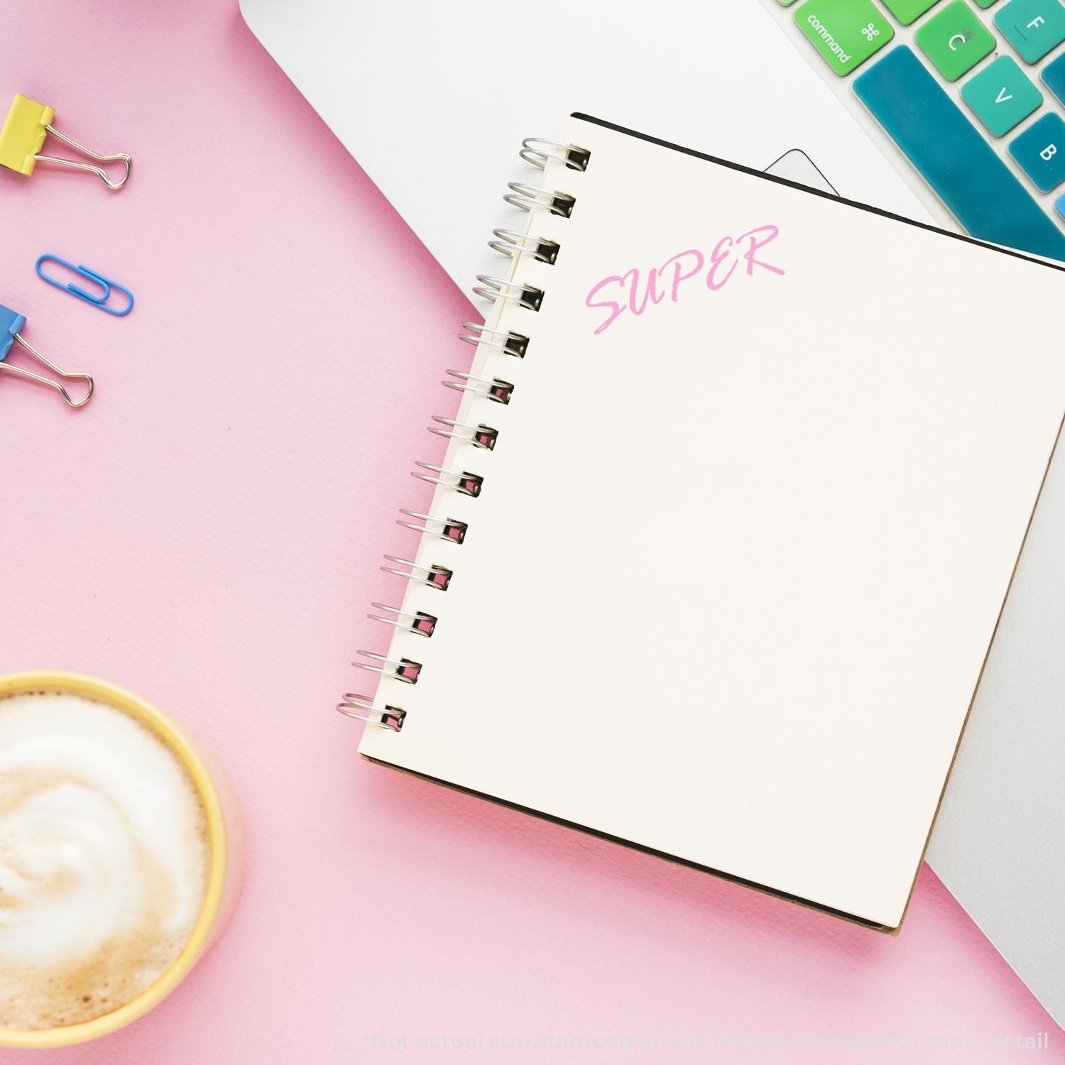 Self Inking Super Stamp imprinting SUPER in pink on a spiral notebook, next to a laptop, coffee cup, and colorful paper clips on a pink desk.