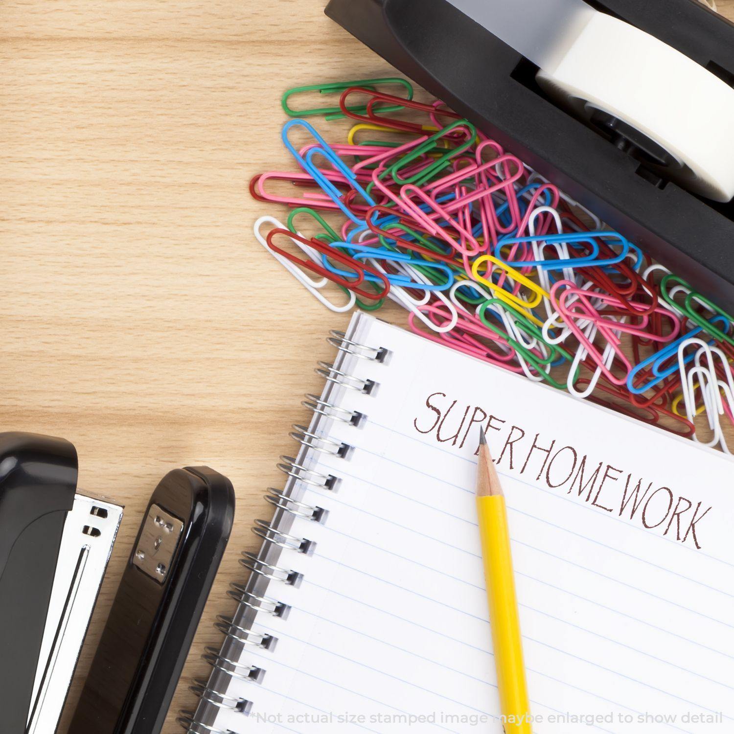 Large Super Homework Rubber Stamp on a notebook page with a pencil, stapler, tape dispenser, and colorful paperclips on a wooden desk.