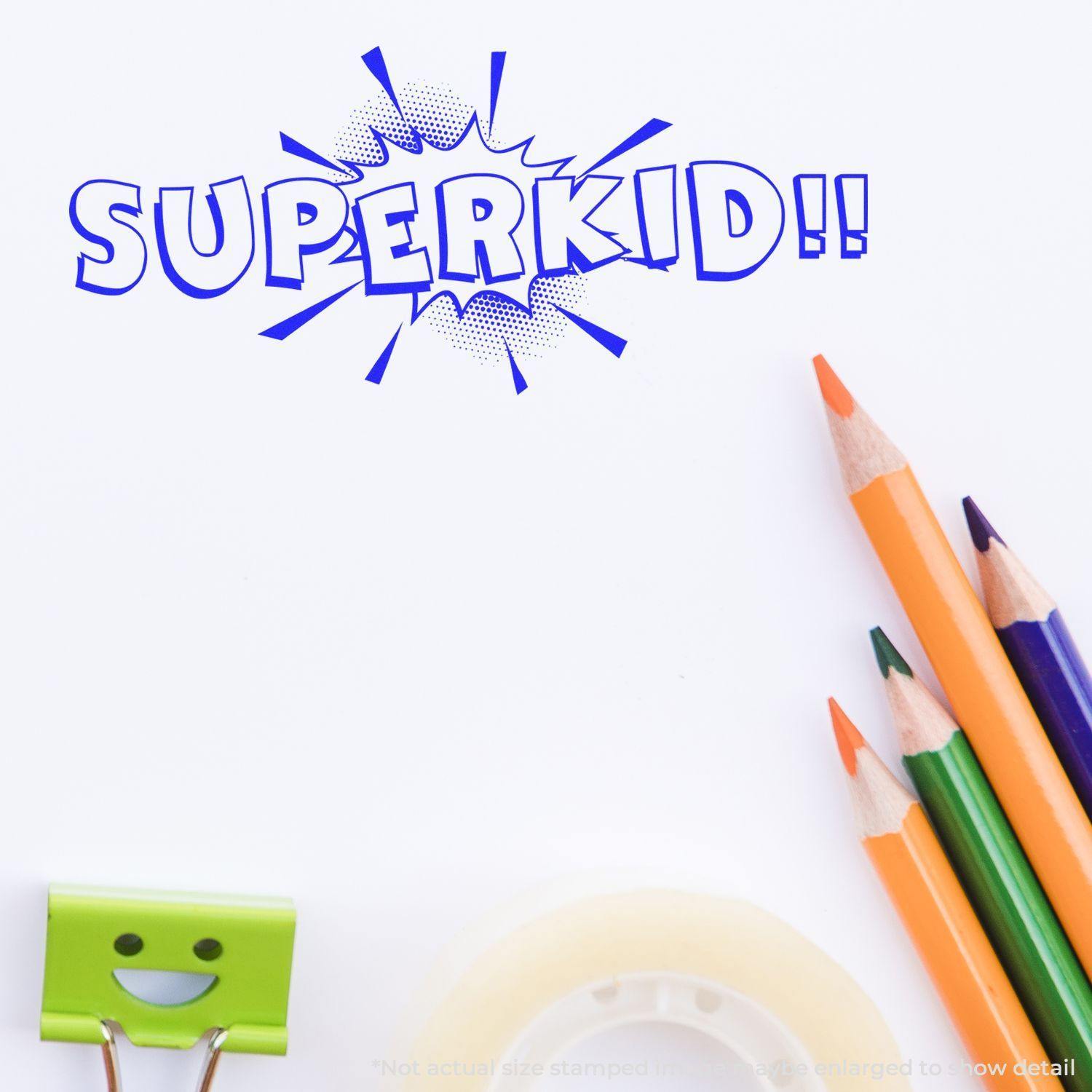 Self Inking Superkid Stamp impression on paper with colorful pencils, a smiling binder clip, and tape in the background.