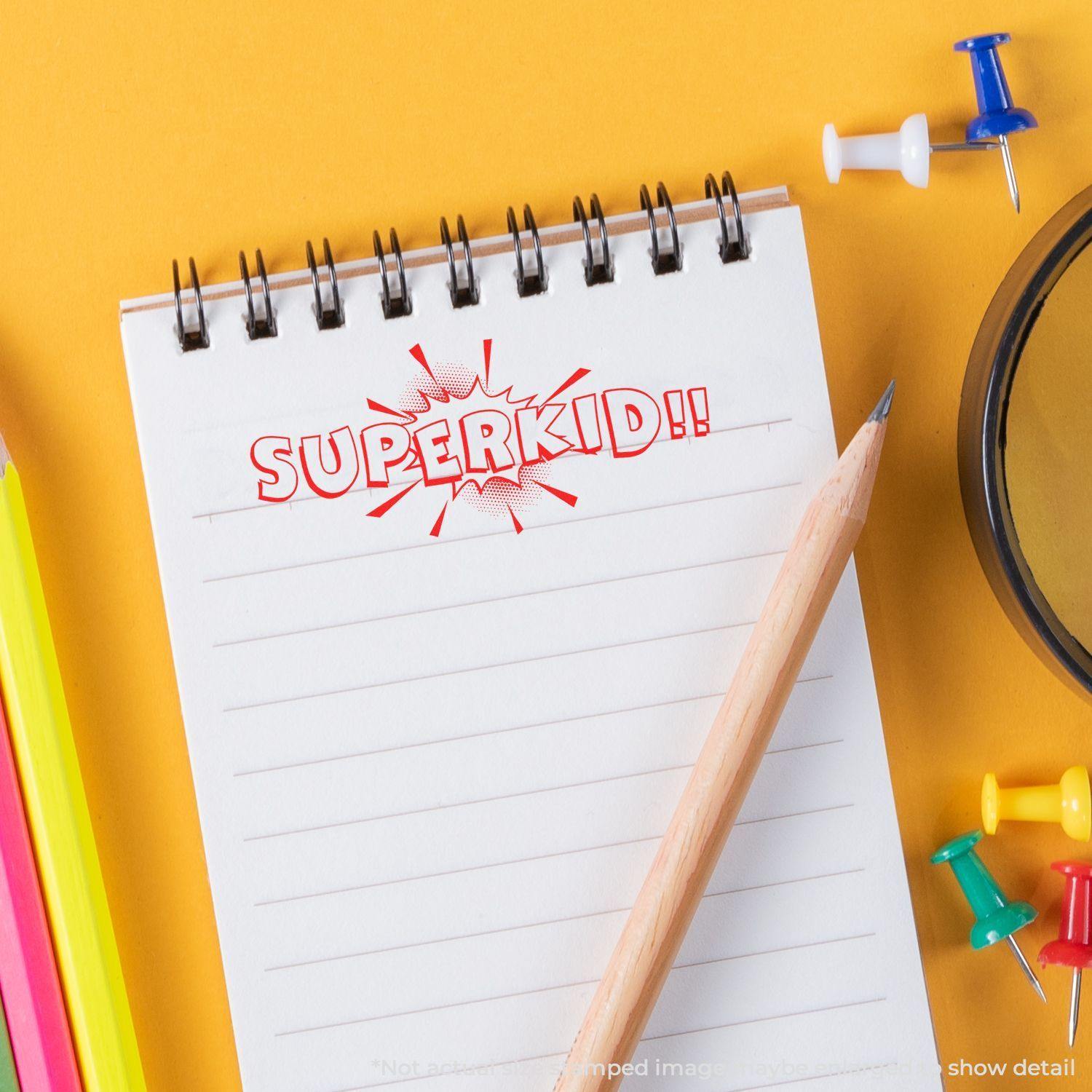 A notebook with a Superkid rubber stamp impression, surrounded by colorful pencils, push pins, and a pencil on a yellow background.