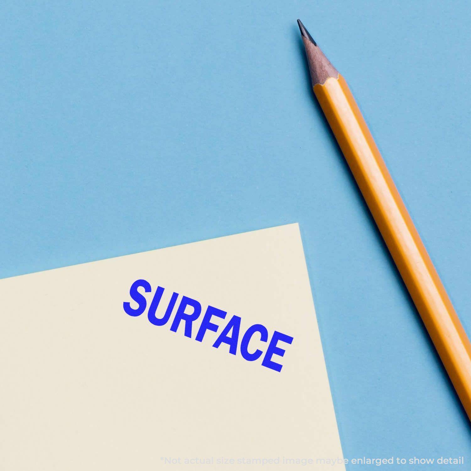 A Large Pre-Inked Surface Stamp imprinting the word SURFACE in blue on a white paper, with a yellow pencil placed beside it.