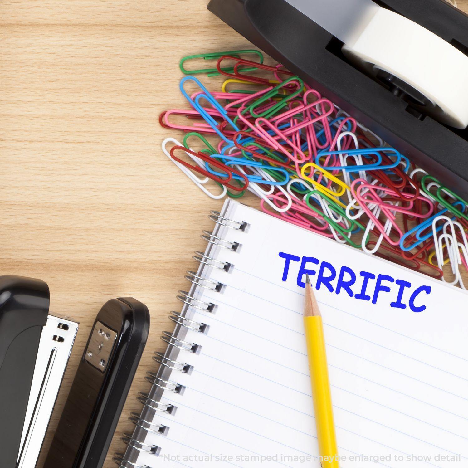 Large Pre-Inked Terrific Stamp used on a notebook page with a pencil, surrounded by colorful paperclips, stapler, and tape dispenser.