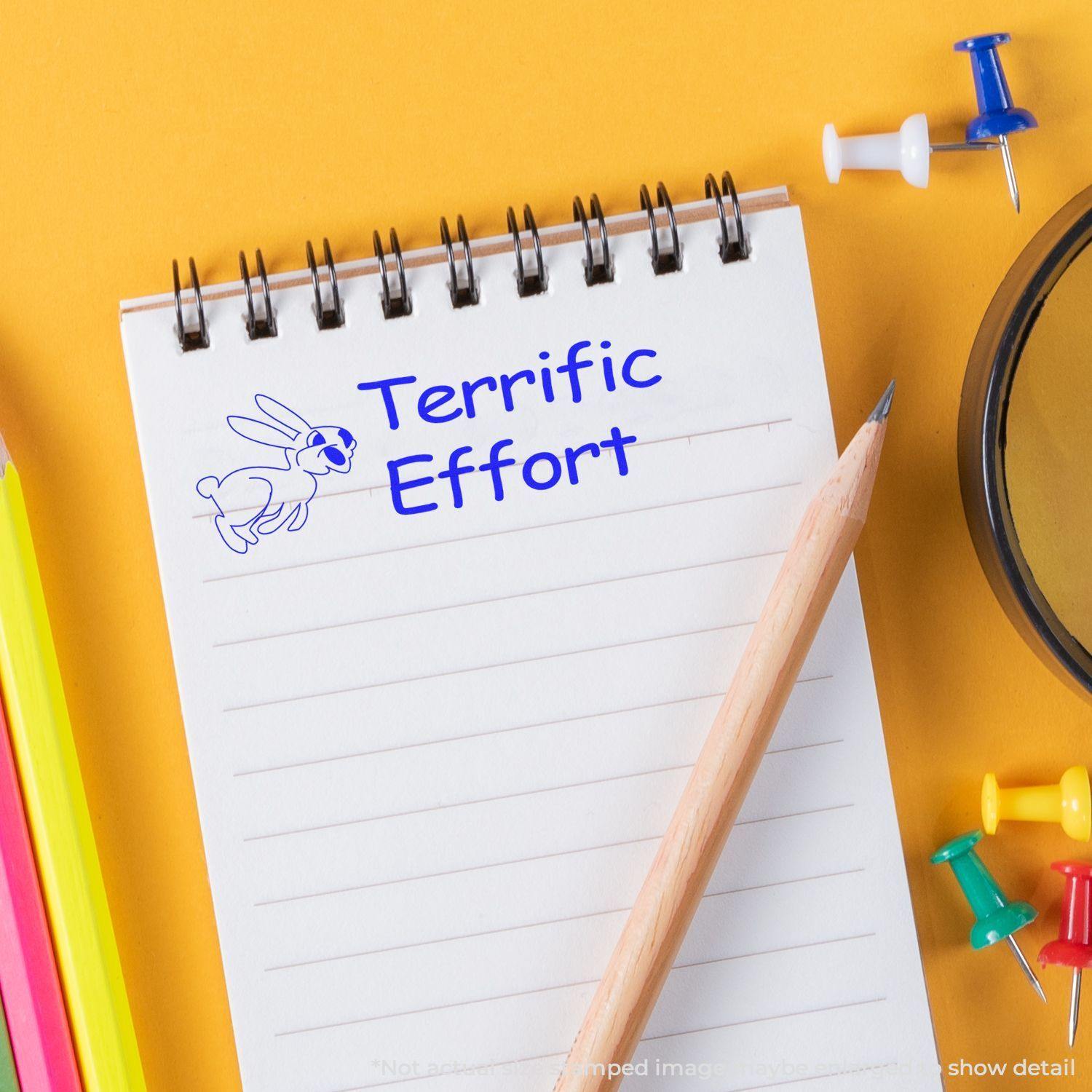 A spiral notebook with a Terrific Effort rubber stamp in blue ink, surrounded by colorful pens, push pins, and a pencil.