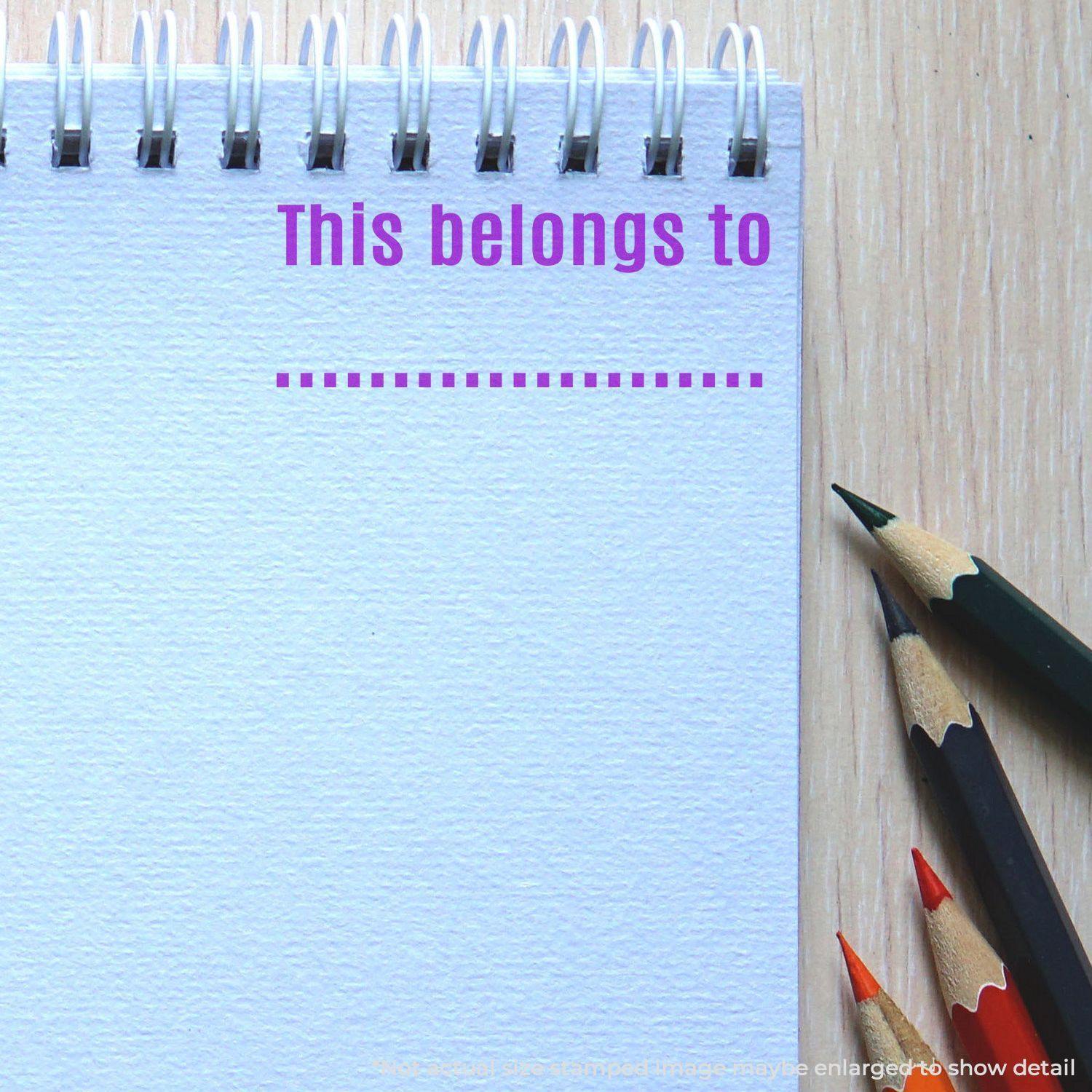 Spiral notebook with This belongs to rubber stamp in purple ink, next to colored pencils on a wooden surface.