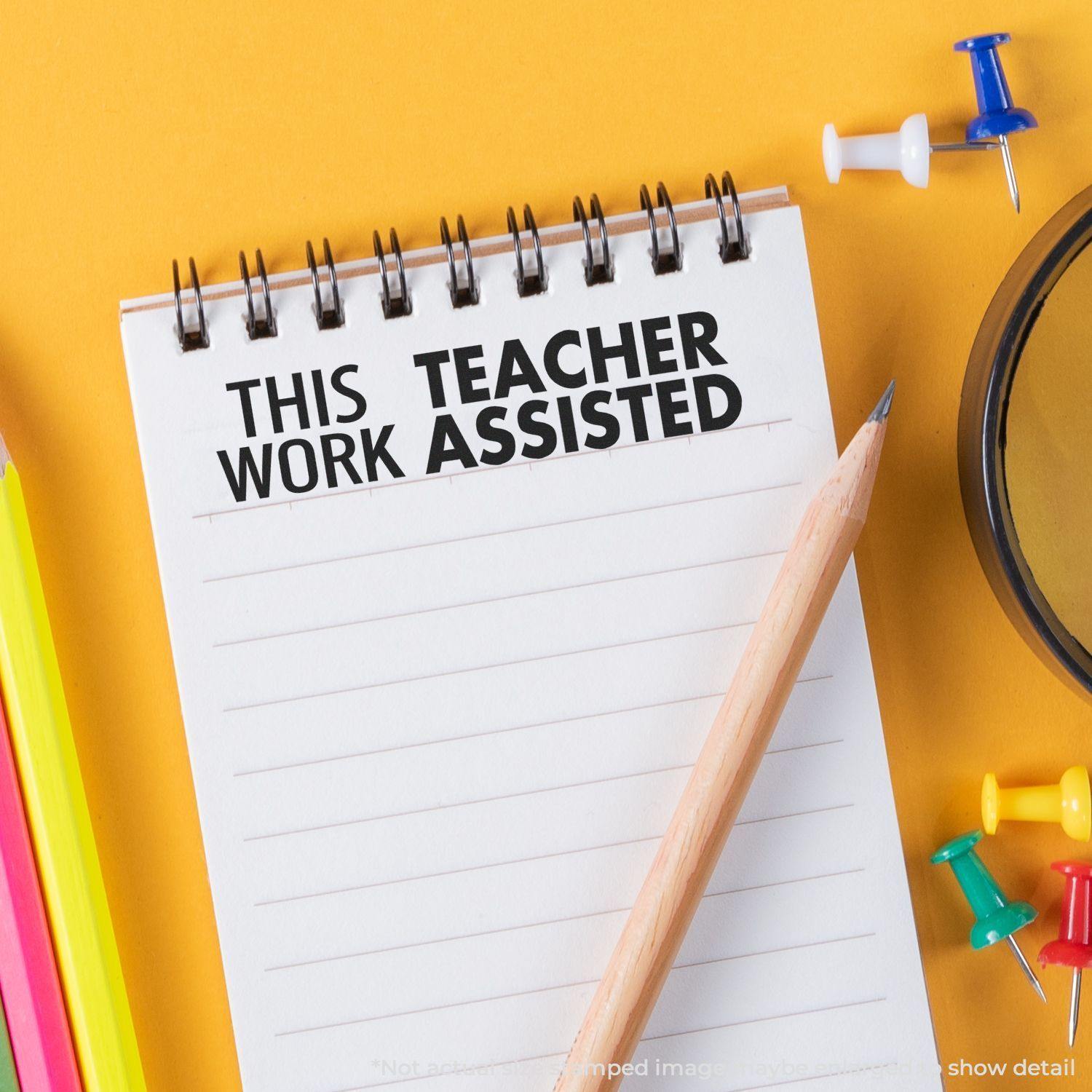 Notepad with This Work Teacher Assisted rubber stamp, surrounded by colorful pencils, push pins, and a pencil on a yellow background.