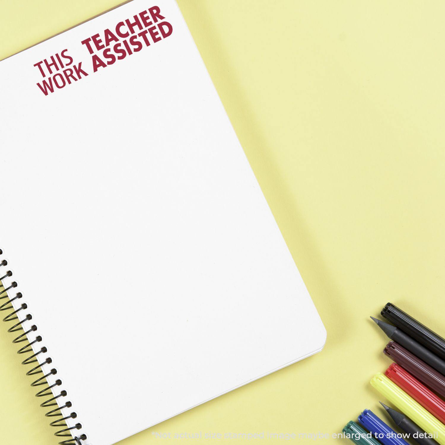 Large This Work Teacher Assisted rubber stamp impression on a spiral notebook, with pens and markers on a yellow background.