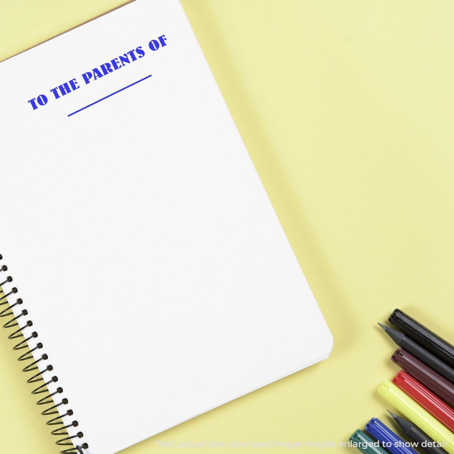 Large Self Inking To The Parents Of Stamp used on a spiral notebook, with colorful pens placed beside it on a yellow background.