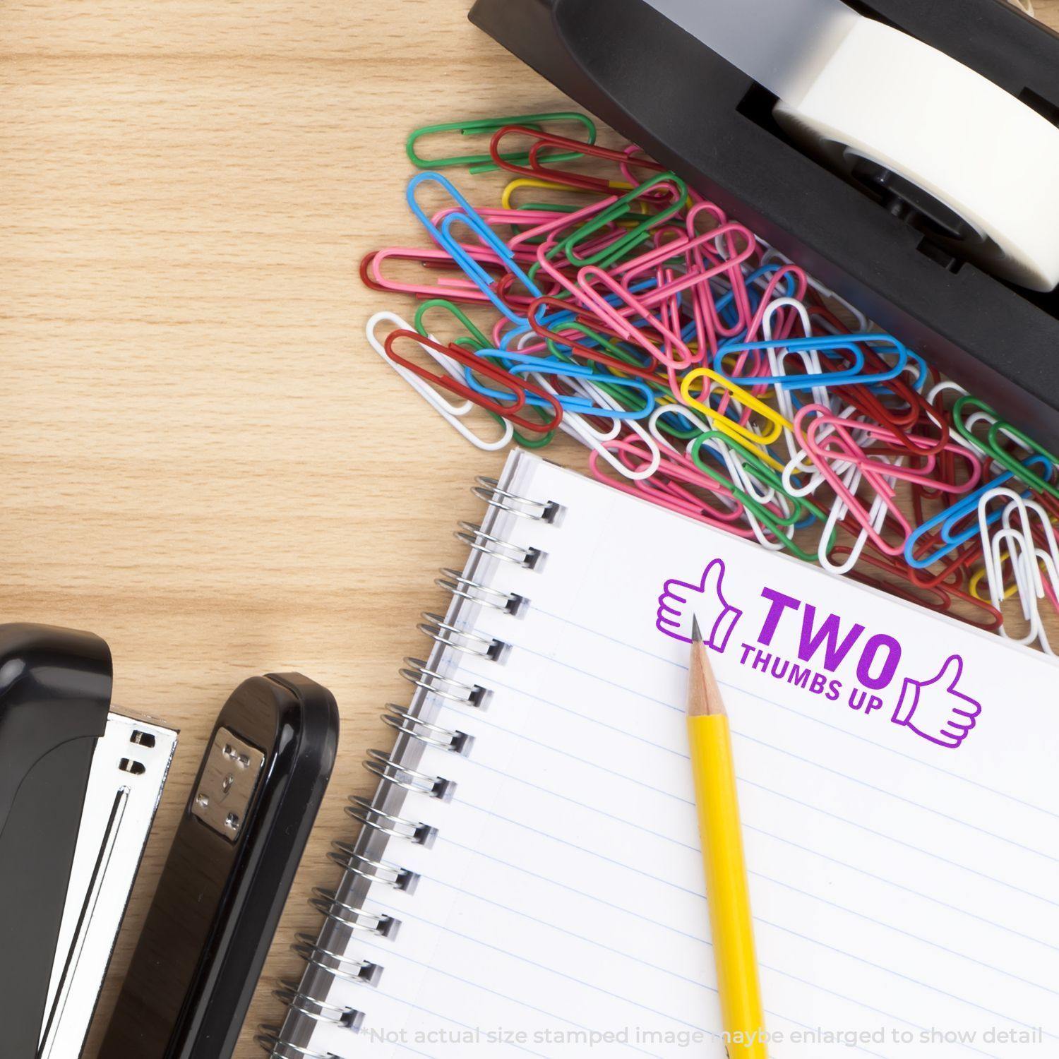 Desk with colorful paperclips, stapler, tape dispenser, and a notebook stamped with Self Inking Two Thumbs Up with Thumb Icon Stamp.