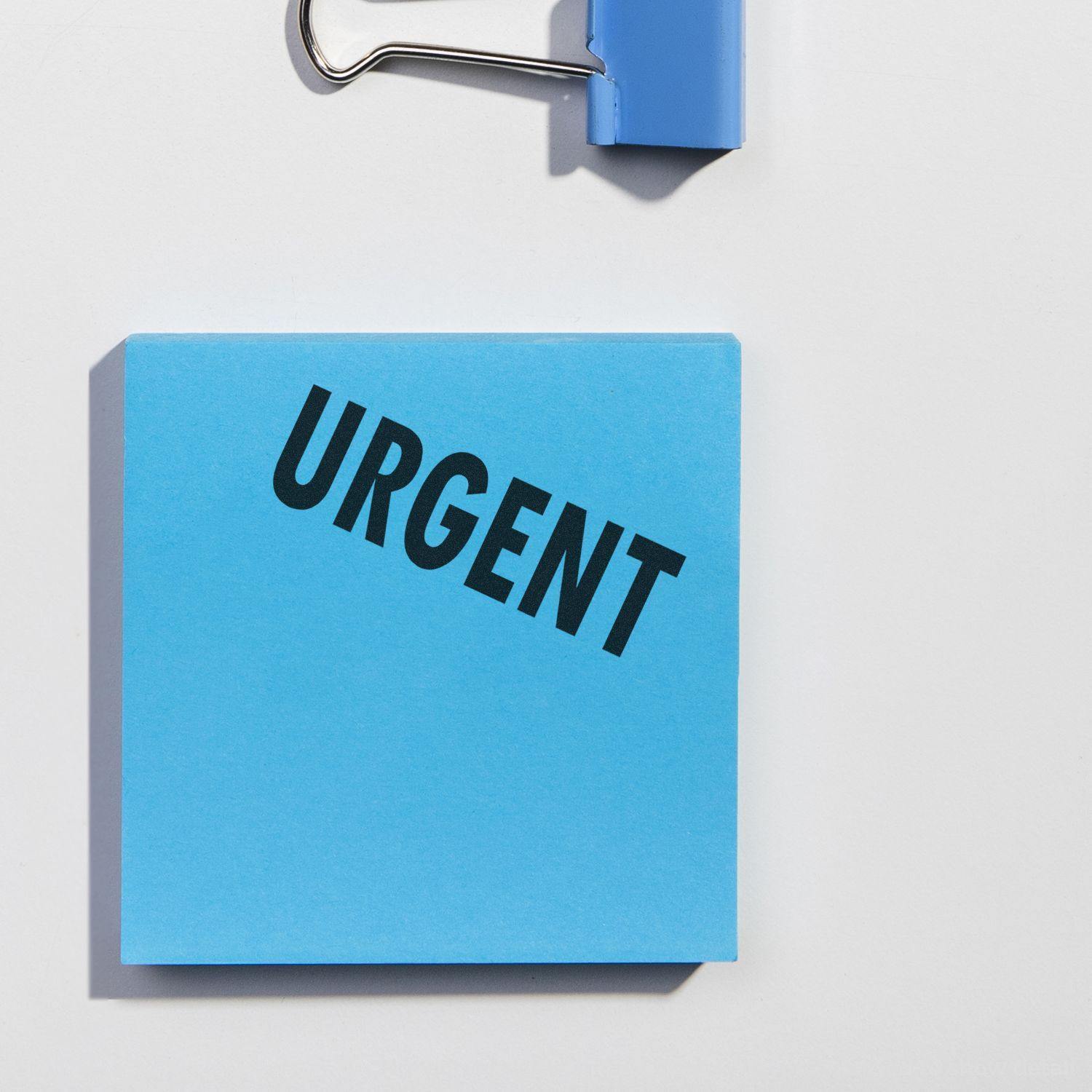 Blue sticky note stamped with URGENT using a Slim Pre-Inked Urgent Stamp, next to a blue binder clip on a white surface.