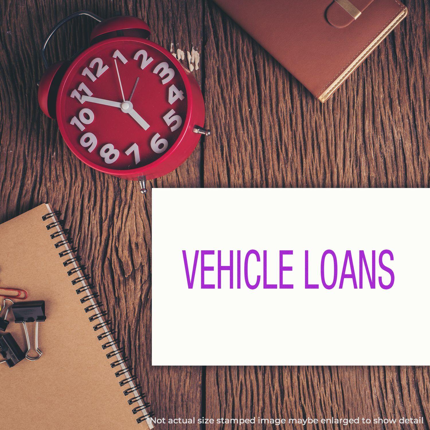 Large Pre-Inked Vehicle Loans Stamp used on a white paper, placed on a wooden desk with a red clock, notebook, and office supplies.