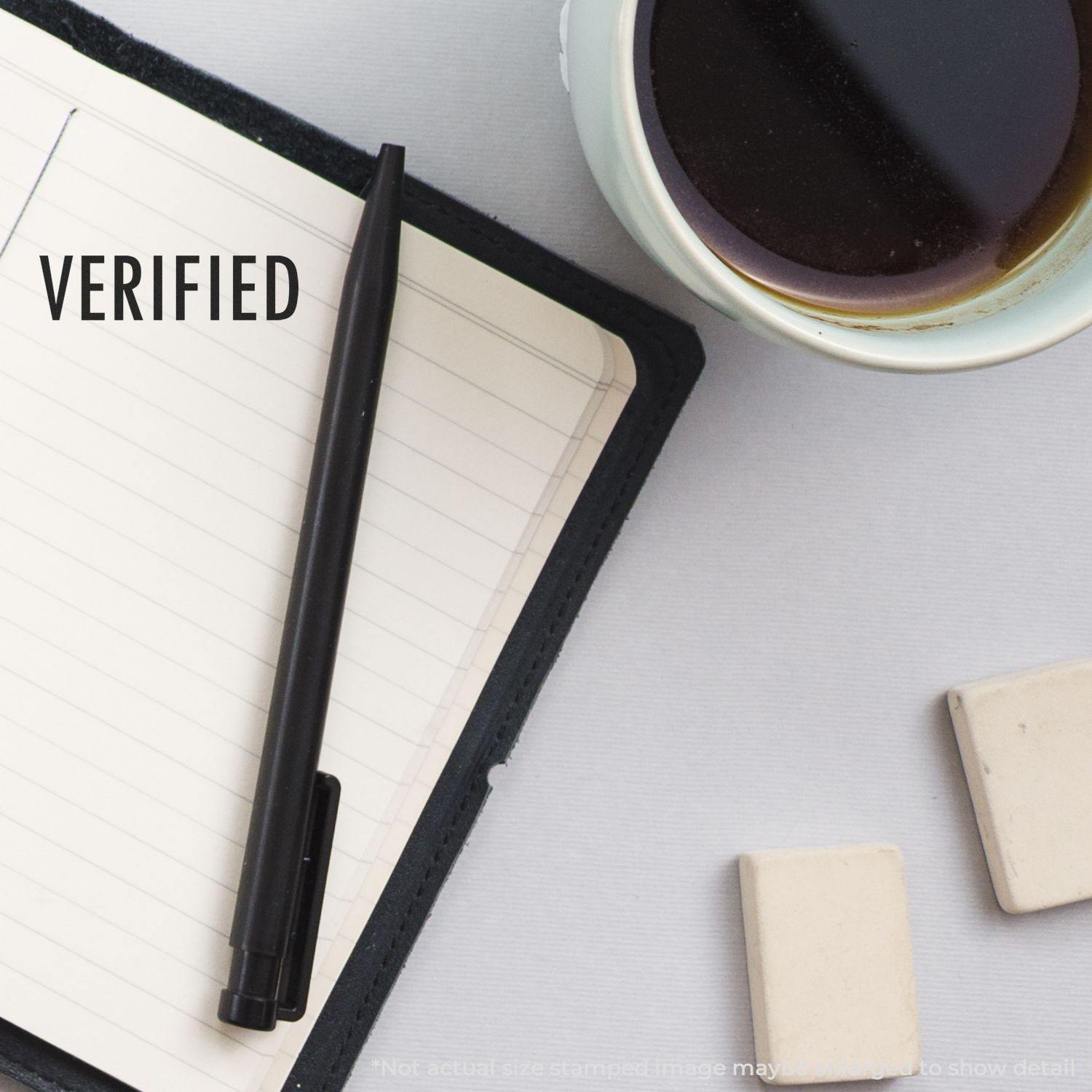 Large verified rubber stamp on a notebook with a black pen, coffee cup, and erasers on a white desk.