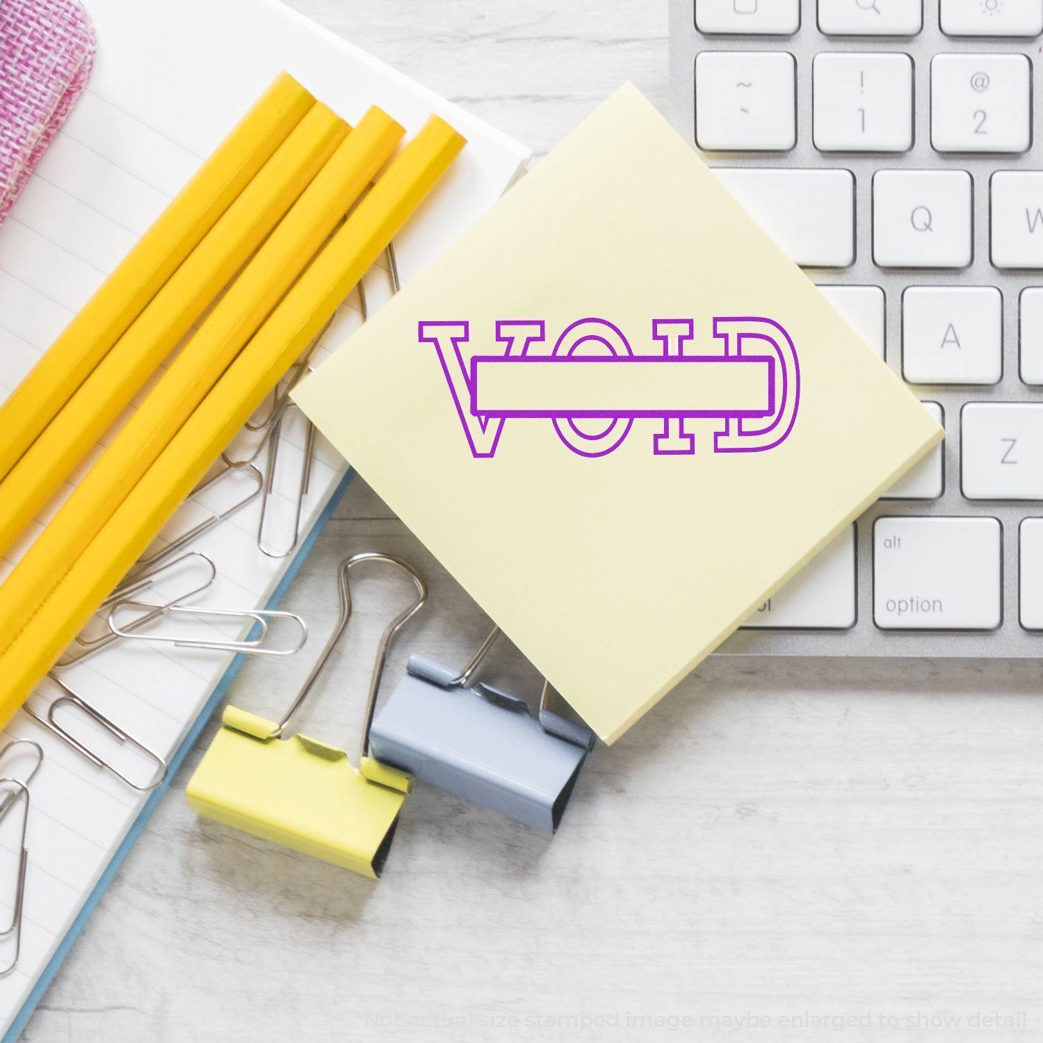 Large Pre-Inked Void with Box Stamp on a sticky note beside a keyboard, pencils, paper clips, and binder clips on a desk.