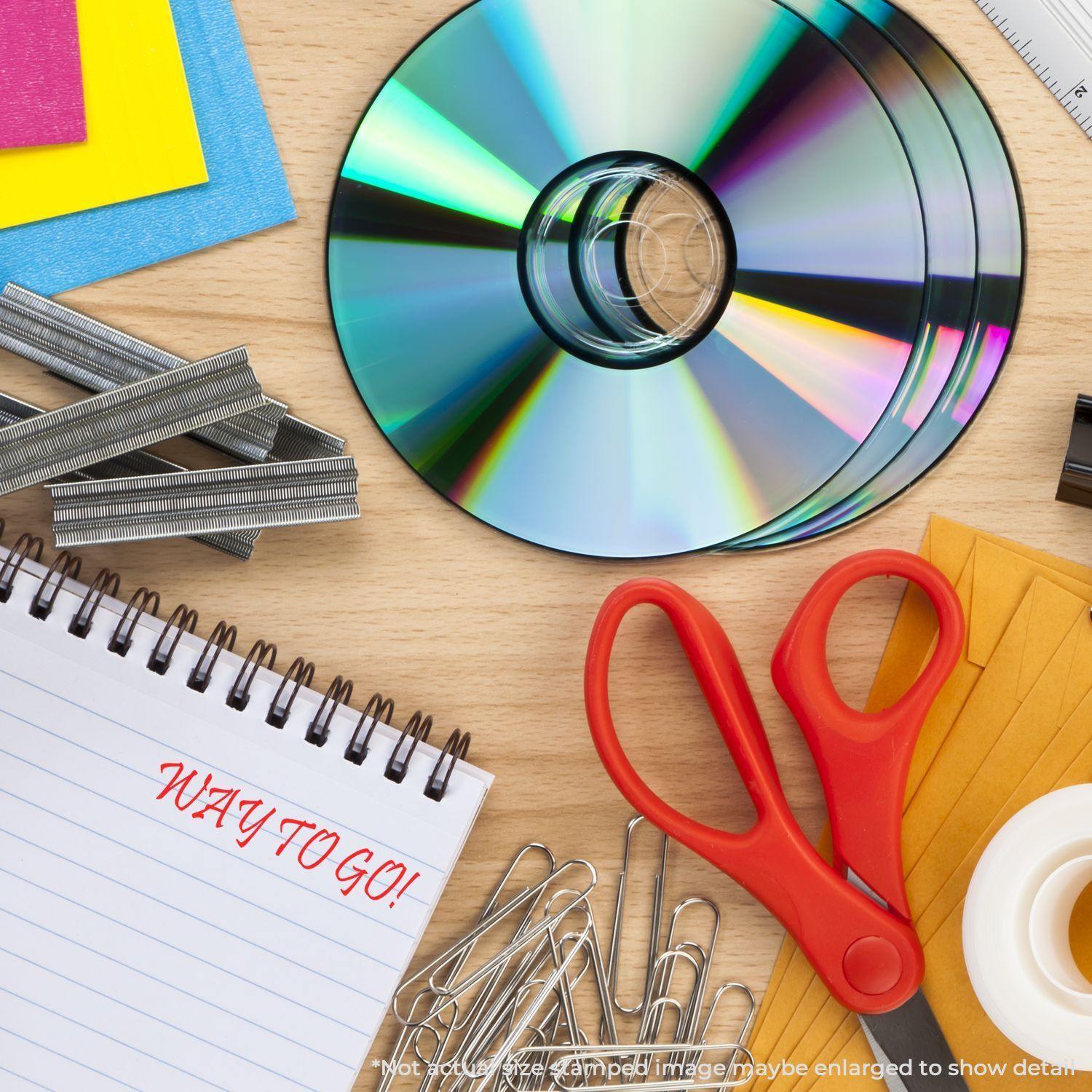 Large Self Inking Way To Go Stamp on a notepad, surrounded by CDs, red scissors, paper clips, staples, and colorful papers on a wooden desk.