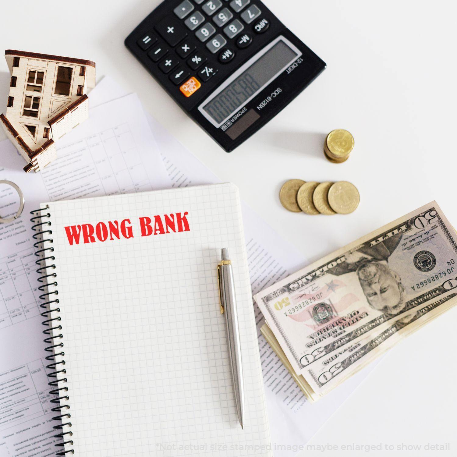 Notebook stamped with WRONG BANK using the Large Pre-Inked Wrong Bank Stamp, surrounded by a calculator, coins, and dollar bills.