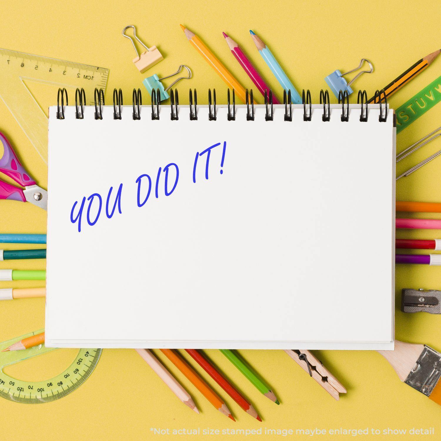 Self Inking You Did It Stamp on a white notebook surrounded by colorful pencils, rulers, and other stationery on a yellow background.