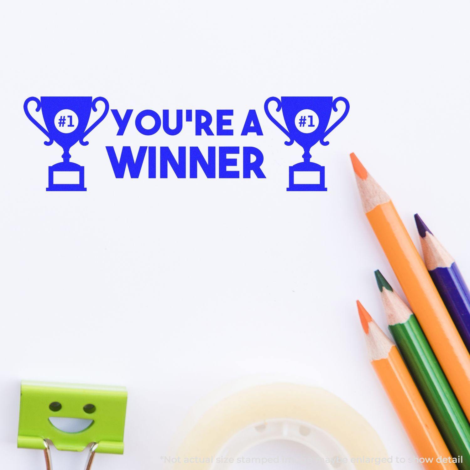 Large Pre-Inked You're a Winner Stamp in blue ink, featuring two trophy icons, with pencils, tape, and a smiling paper clip nearby.