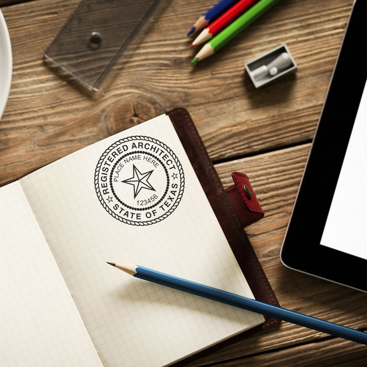 Texas Architect Seal Stamp on an open notebook, surrounded by a pencil, colored pencils, a sharpener, and a tablet on a wooden desk.