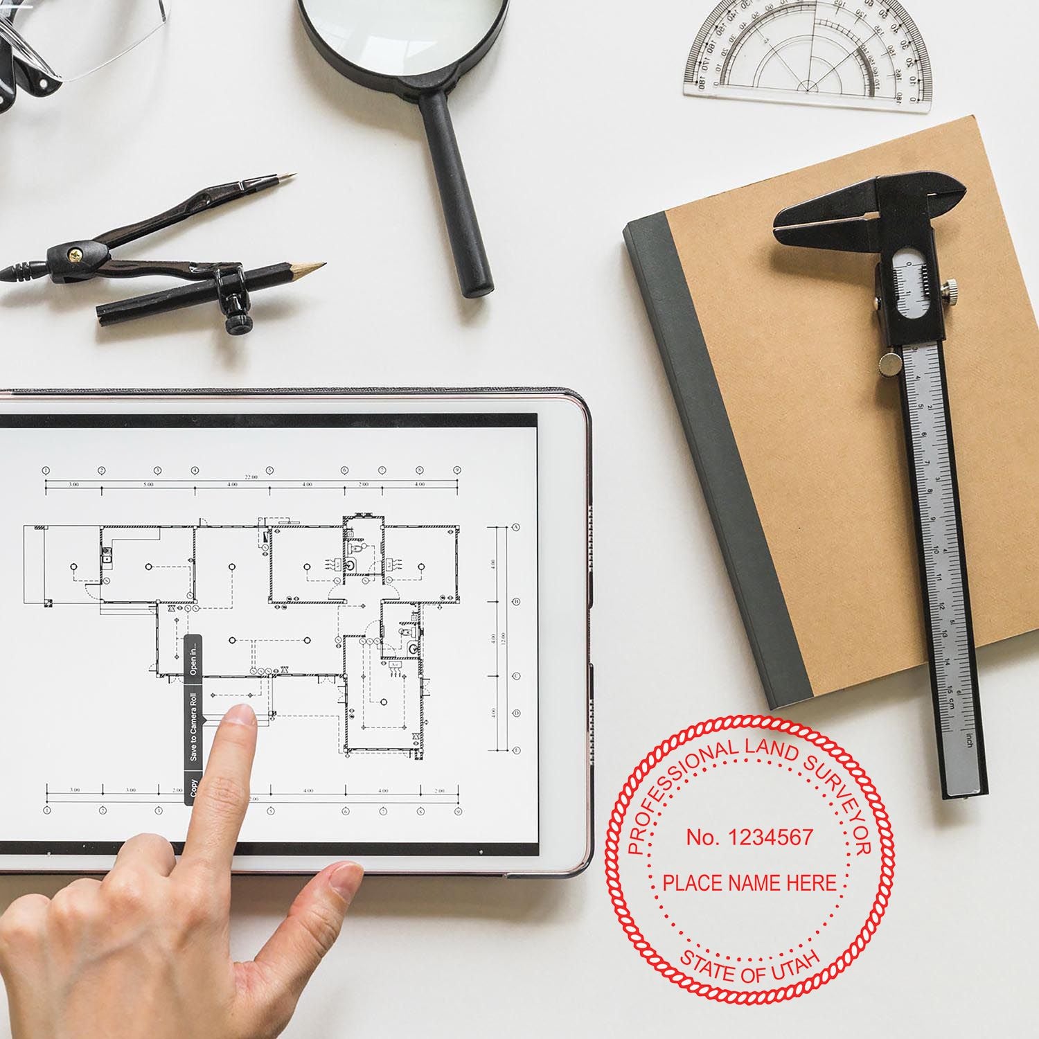 Self Inking Utah Land Surveyor Stamp in use on a desk with architectural tools, a tablet displaying a floor plan, and a notebook.