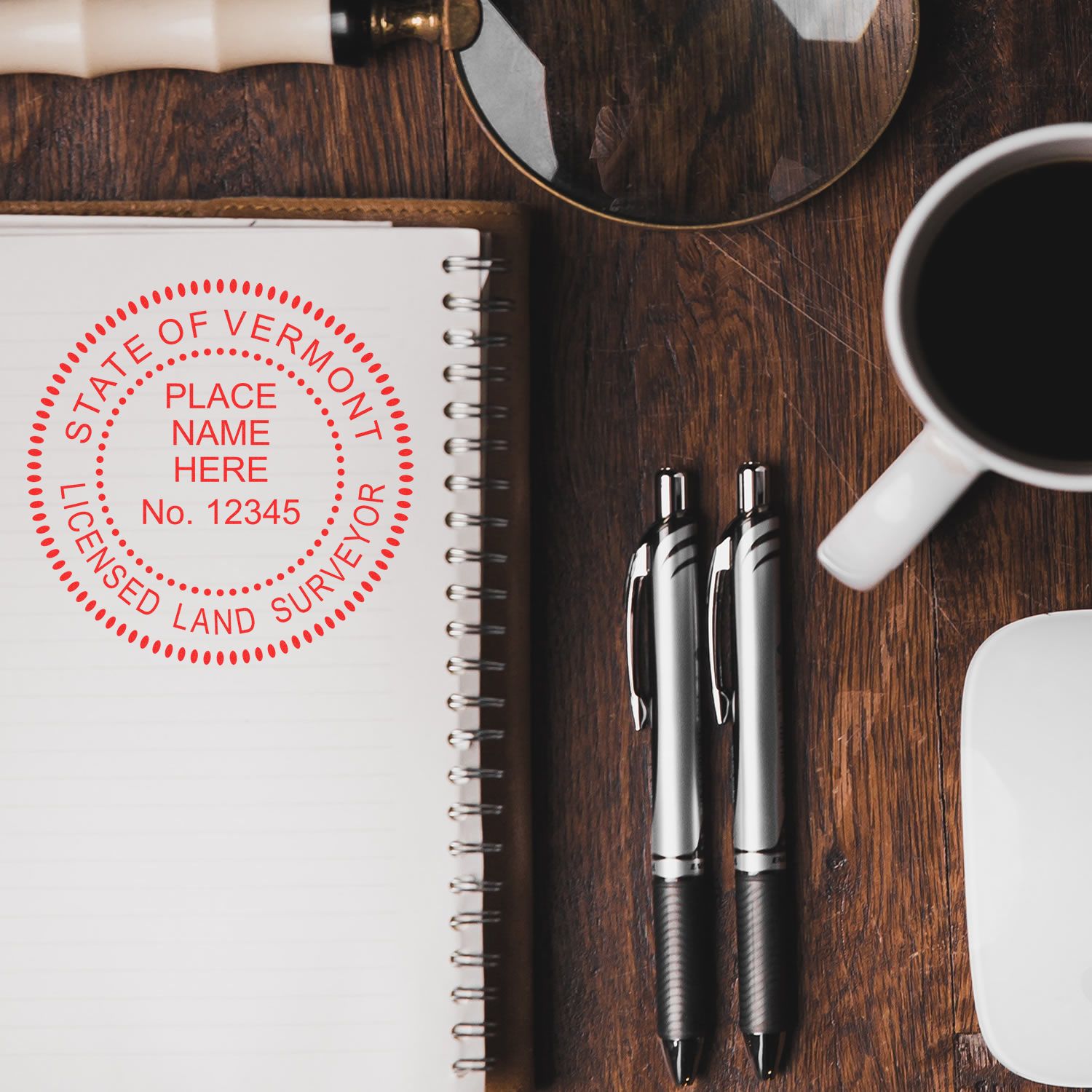 Self Inking Vermont Land Surveyor Stamp on a notebook with pens, magnifying glass, and coffee cup on a wooden desk.