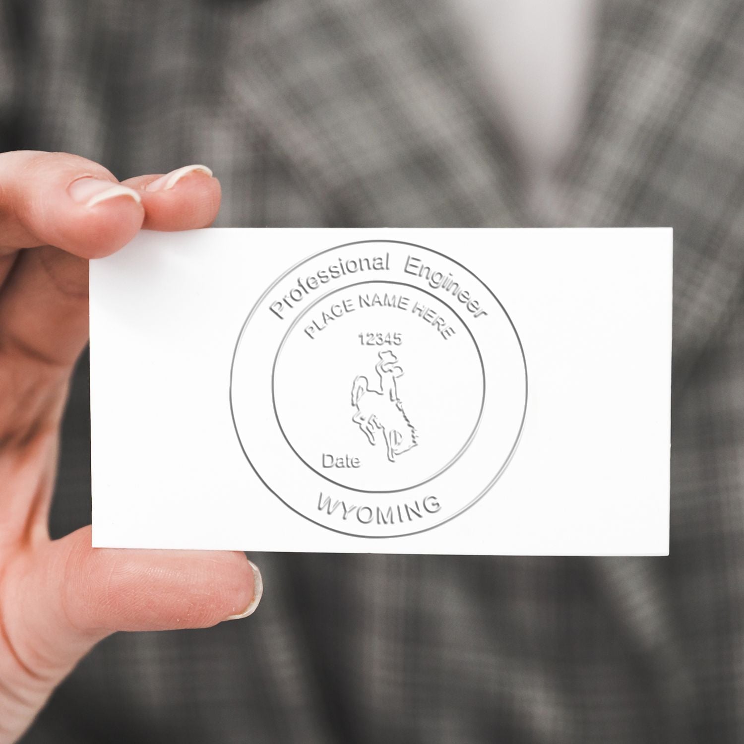 A hand holding a white card embossed with a Wyoming seal using the Professional Engineer Hybrid Seal Embosser. The seal includes text Professional Engineer, Place Name Here, Date, and WYOMING.