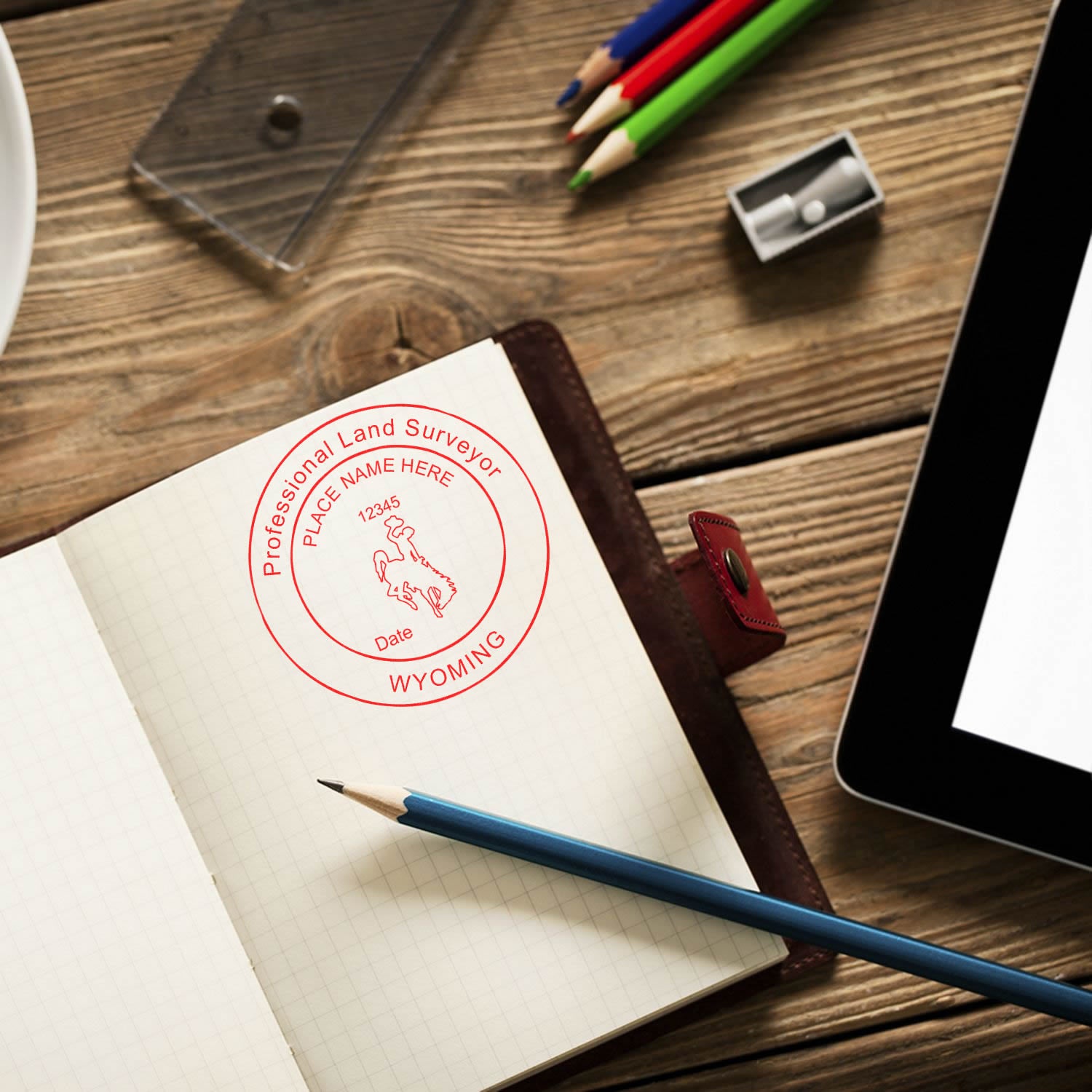 Self Inking Wyoming Land Surveyor Stamp in use on a notebook, surrounded by pencils, a sharpener, and a tablet on a wooden desk.