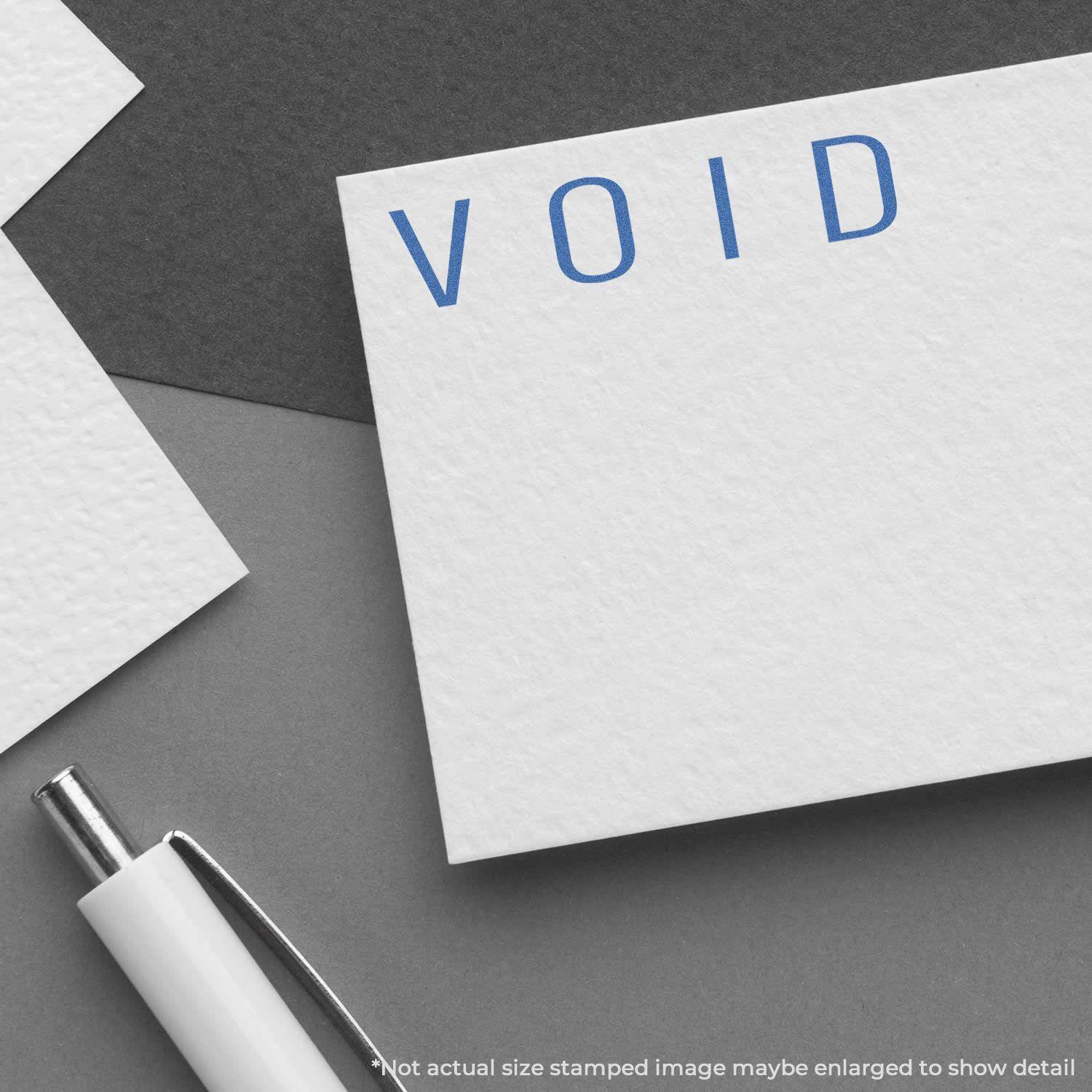 A Blue Void Xstamper Stamp imprint on white paper, showing the word VOID in blue ink, with a pen and additional papers nearby.